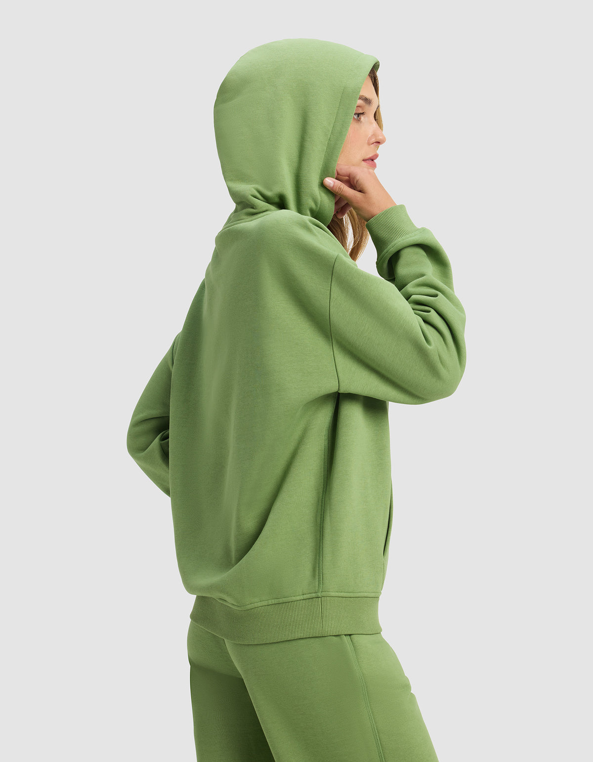 A woman in profile wears the Cozy Earth Women's Cityscape Hoodie and matching sweatpants in green, standing against a plain light background with her hood up and one hand touching her chin.
