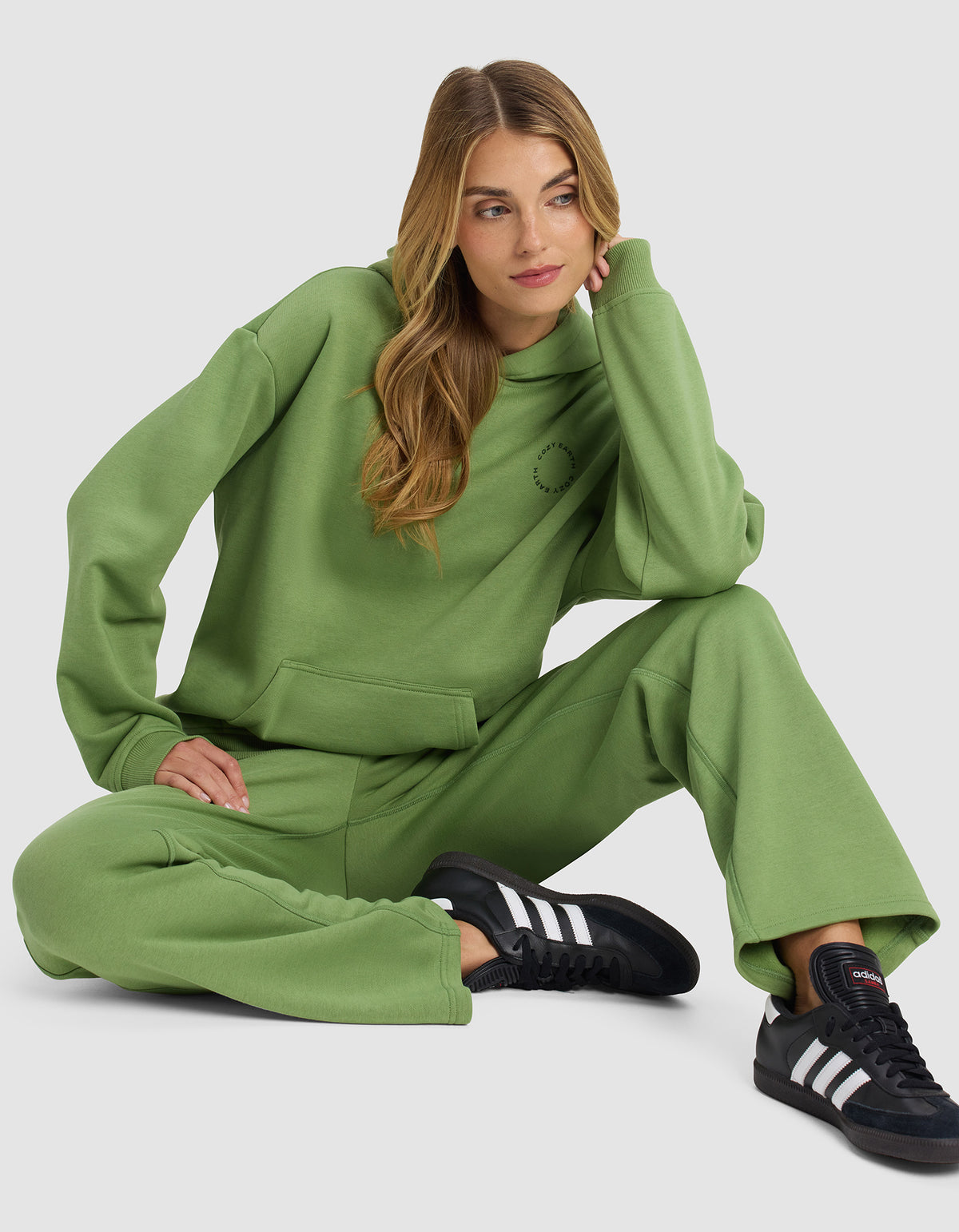 A woman with long, wavy hair sits on the floor wearing Cozy Earth’s Women’s Cityscape Hoodie in green and matching sweatpants, paired with black Adidas sneakers. She rests her head on one hand and looks to the side.