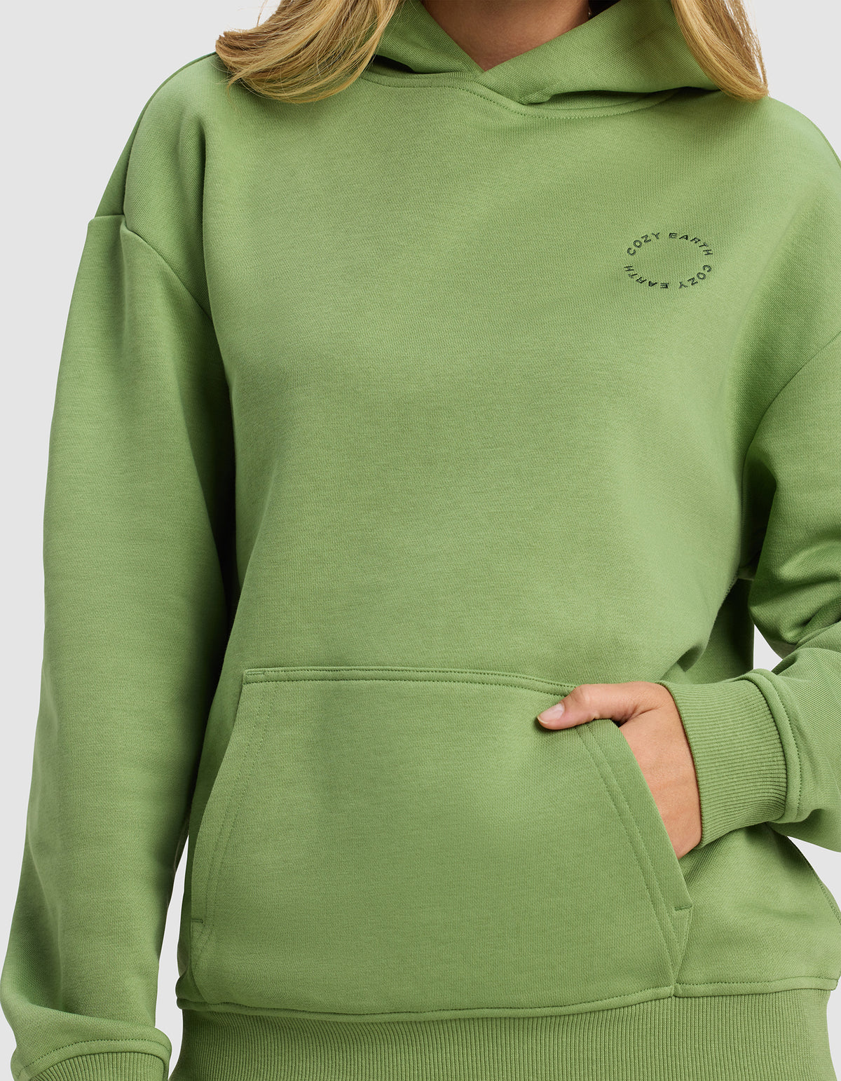 A woman wears the Cozy Earth Women's Cityscape Hoodie in light green, featuring a front pocket and small circular "COZY EARTH" text on the chest. Only her lower face, neck, and upper body are visible.