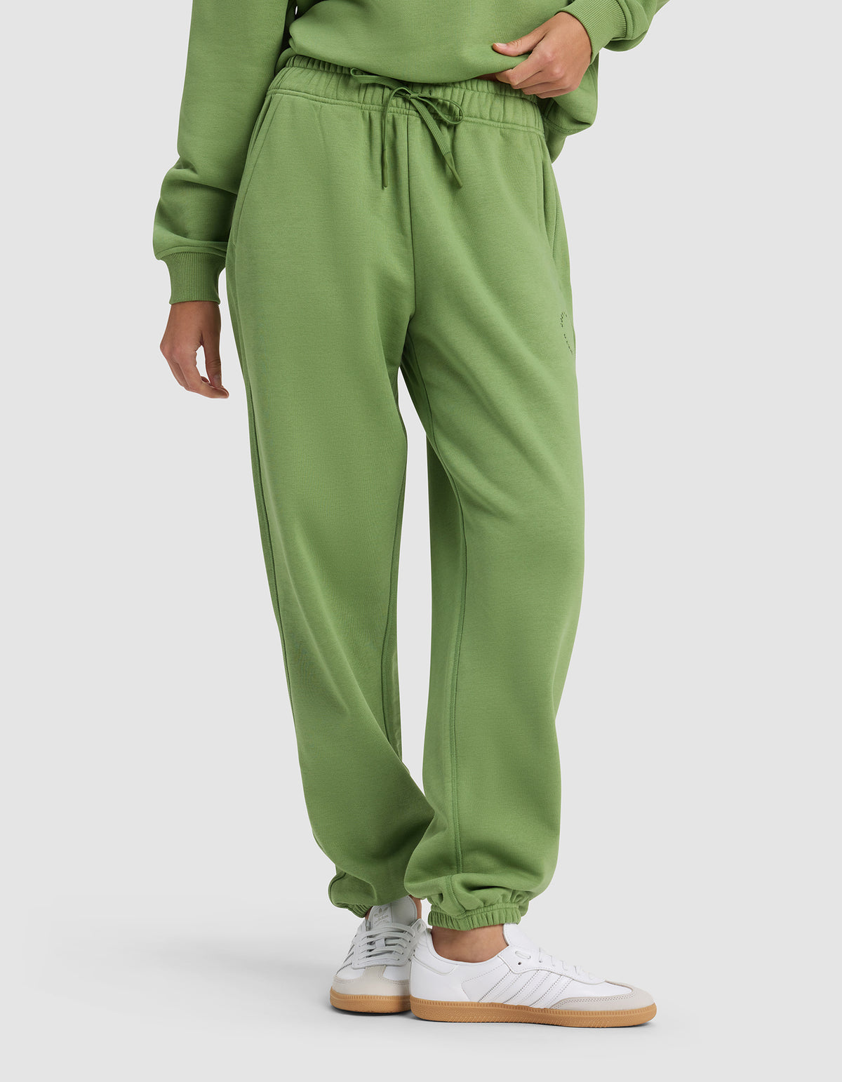 A person wears Cozy Earth's Women's Cityscape Sweatpant—a green pair with an elastic waistband and drawstring—styled with a matching sweatshirt and white sneakers, standing against a plain light background.