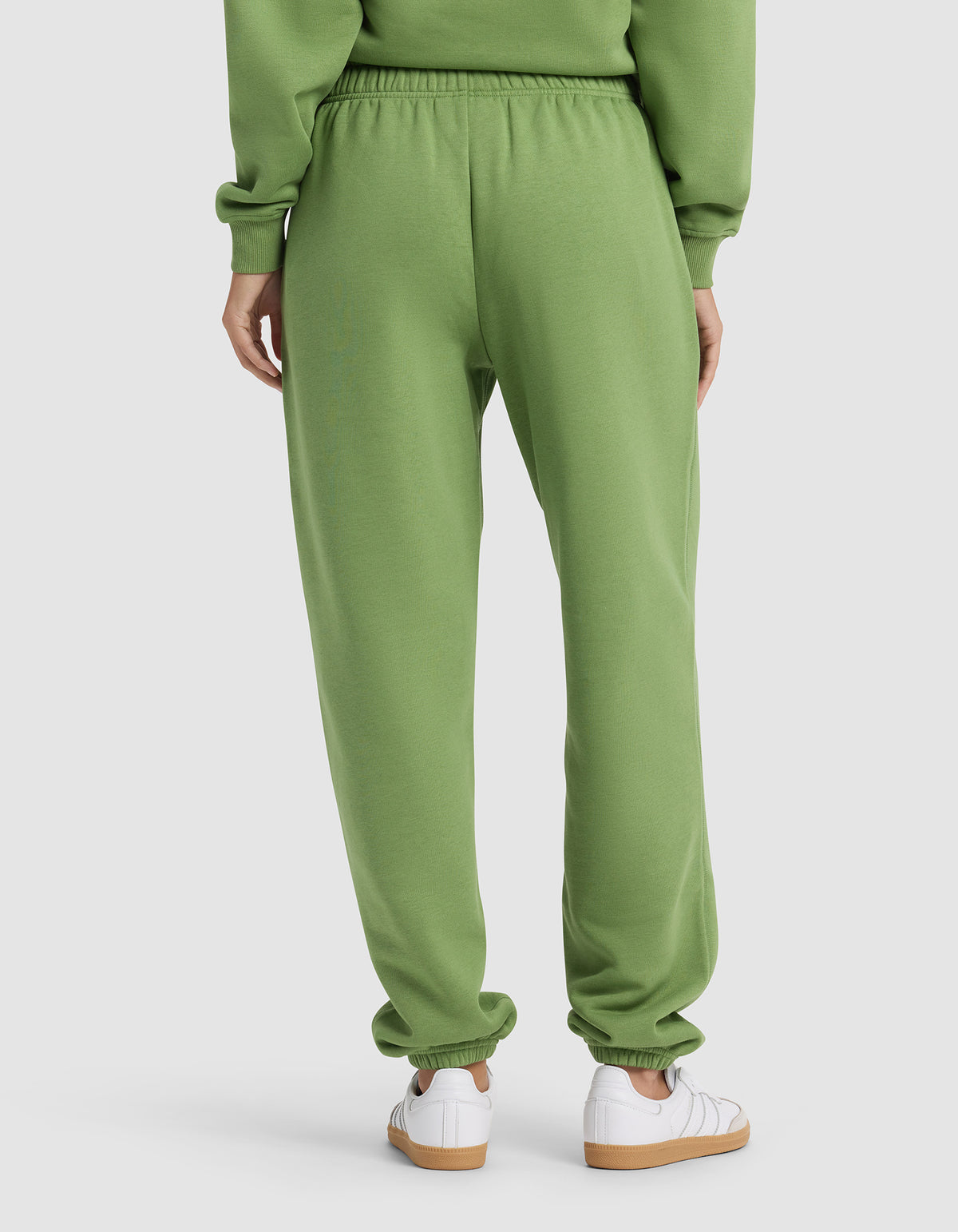 A woman is seen from behind in Cozy Earth's Women's Cityscape Sweatpant and a matching green sweatshirt, paired with white sneakers, against a plain background.