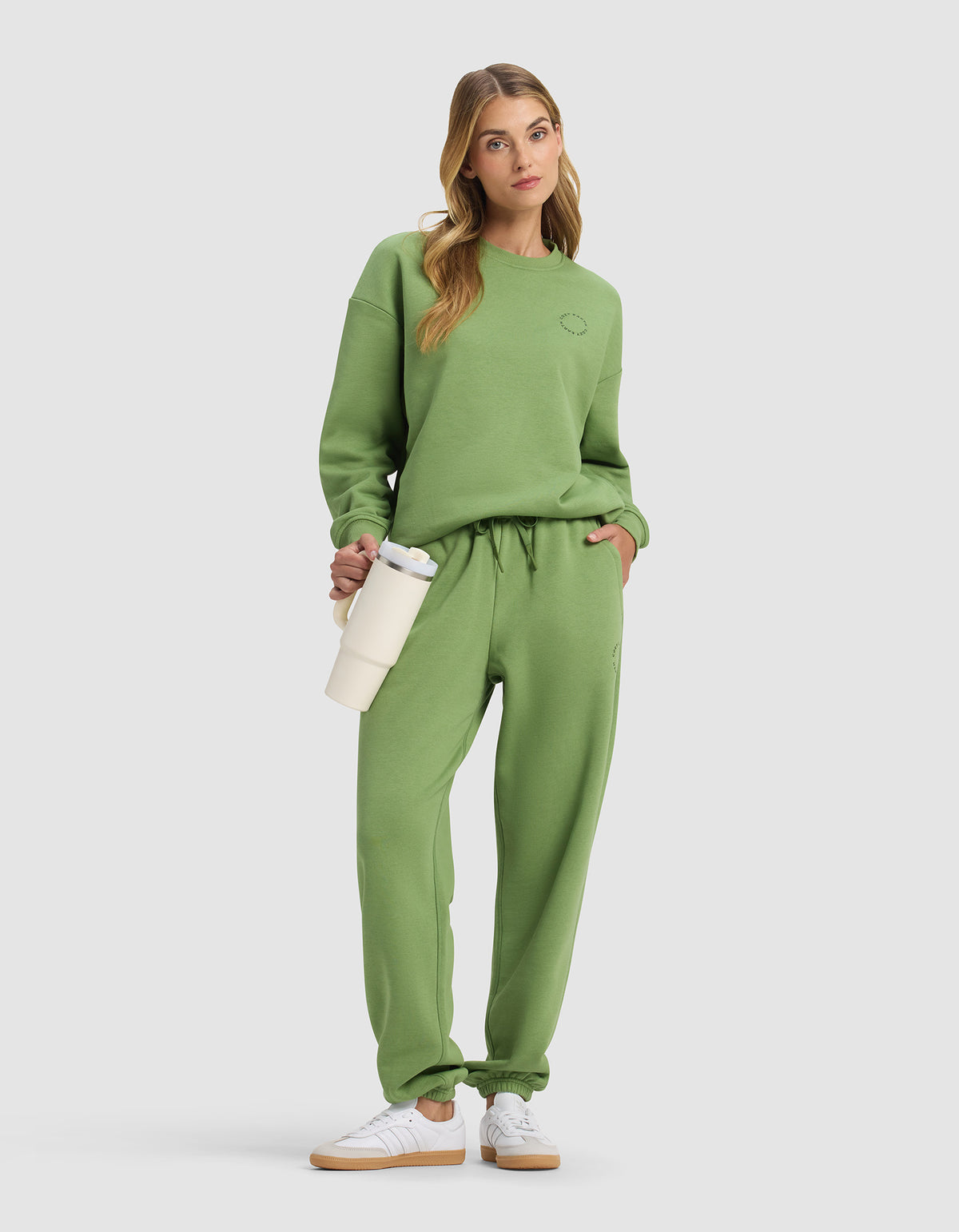 A woman models the Cozy Earth Women's Cityscape Sweatpant in green with a matching sweatshirt, white sneakers, and a cream water bottle, posing with one hand in her pocket against a plain light background.