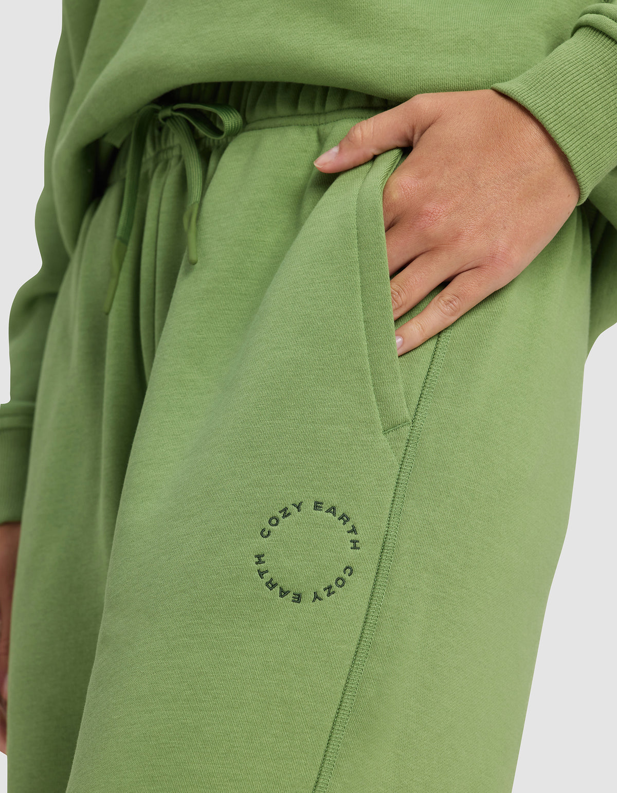 A person wears Cozy Earth's Women's Cityscape Sweatpant in light green with a matching sweatshirt, one hand in their pocket. "COZY EARTH" is printed in a circle near the sweatpant’s pocket.