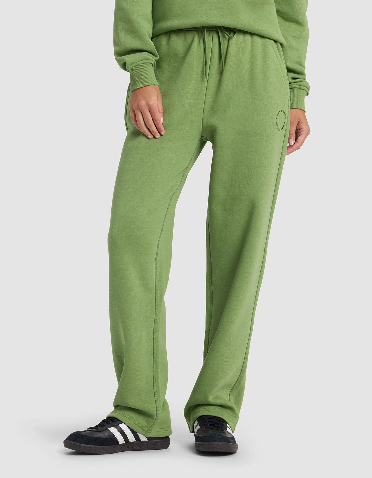 A woman wears Cozy Earth’s Women’s Cityscape Wide Leg Pant in green with a matching sweatshirt and black sneakers with white stripes, standing against a plain white background.
