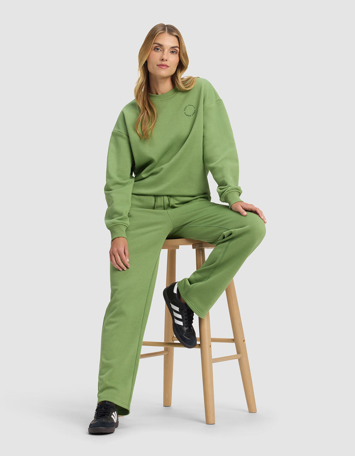 A woman with long blond hair sits on a wooden stool, wearing Cozy Earth's Women's Cityscape Wide Leg Pant in green with a matching loose sweatshirt and black Adidas sneakers against a plain light gray background.