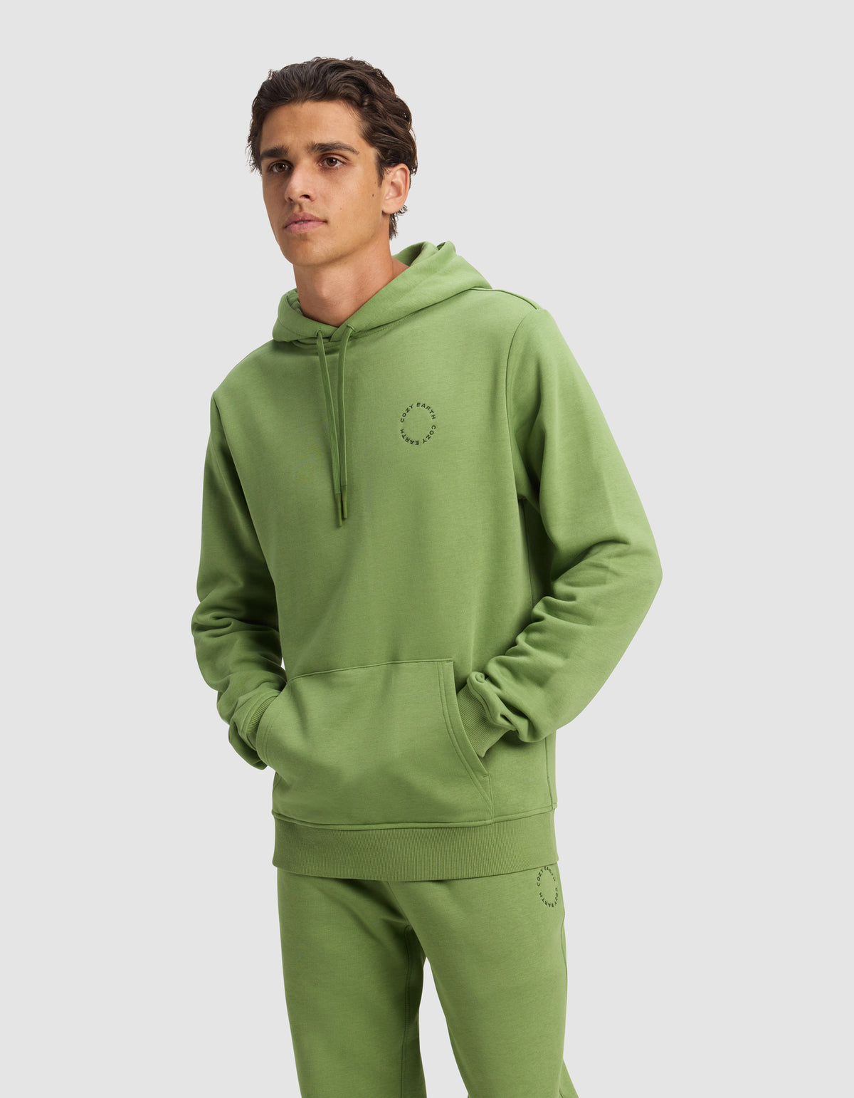 A man wears a Cozy Earth Men's Cityscape Hoodie and matching sweatpants in Clover, both featuring a small circular stitched design, with hands in the hoodie pocket against a plain light gray background.