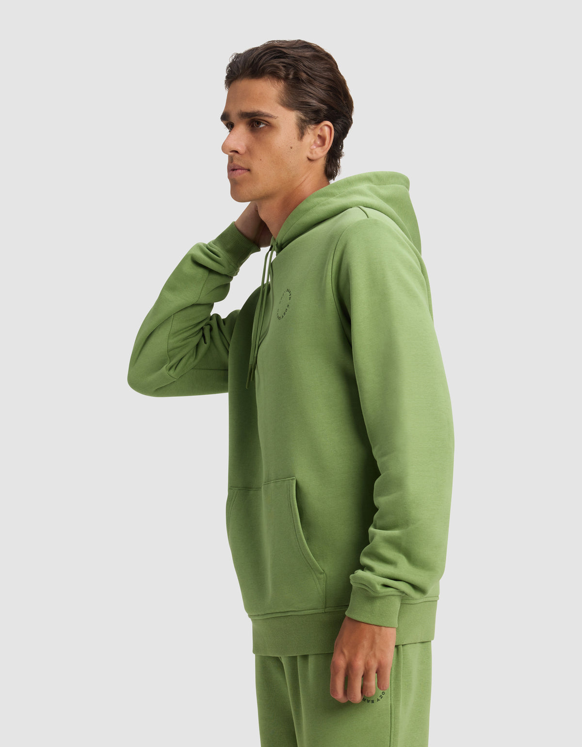 A young man in profile wears the Cozy Earth Men's Cityscape Hoodie in light green, paired with matching joggers. His left hand touches the back of his neck against a plain light gray background.