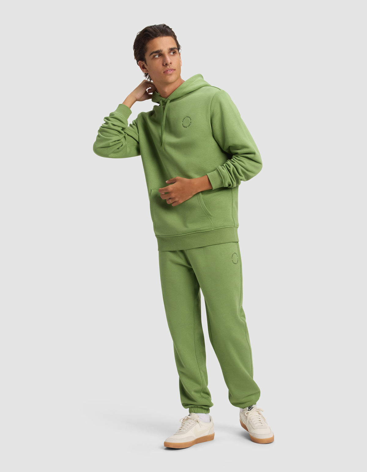 A young man in the Cozy Earth Men's Cityscape Hoodie and matching sweatpants with white sneakers stands against a plain background, looking to the side with one hand touching his neck.