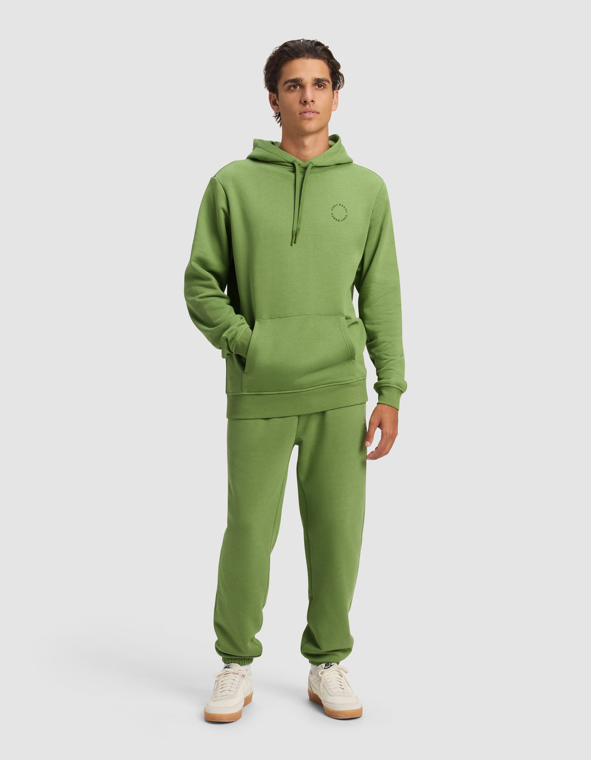 A young man wears the Cozy Earth Men's Cityscape Hoodie & Sweatpant Set with white sneakers, standing against a plain background with his hands in the hoodie pocket and a neutral expression.