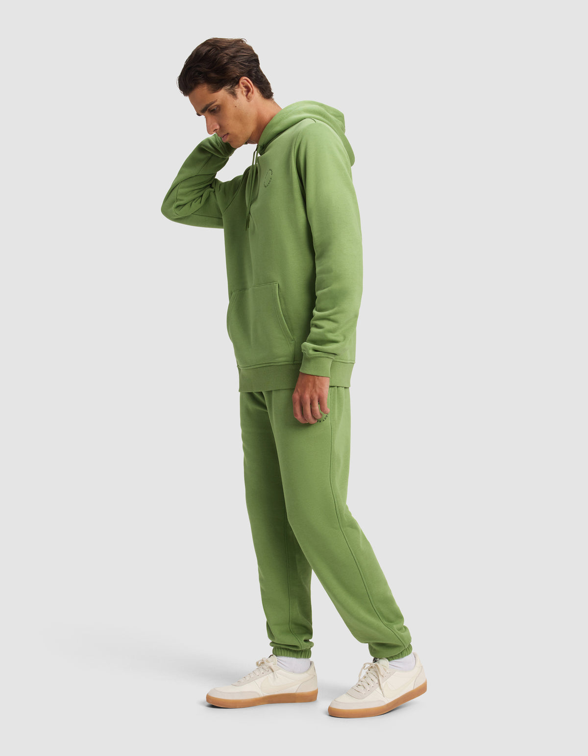 A man wears the Cozy Earth Men's Cityscape Hoodie & Sweatpant Set in light green, paired with white sneakers with tan soles, standing against a plain light background and touching the back of his neck.
