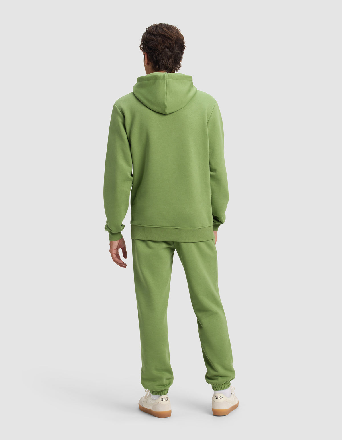 A person with short brown hair is standing and facing away, wearing Cozy Earth's Men's Cityscape Hoodie & Sweatpant Set in light green and white sneakers with beige soles, against a plain light background.