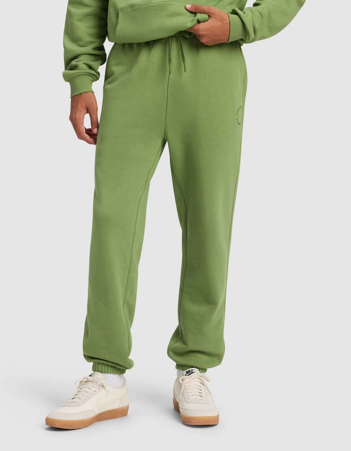 A person wears Cozy Earth's Men's Cityscape Sweatpant in green, paired with a matching hoodie and white-beige Nike sneakers, standing against a plain light background. Only the lower half of their body is visible.