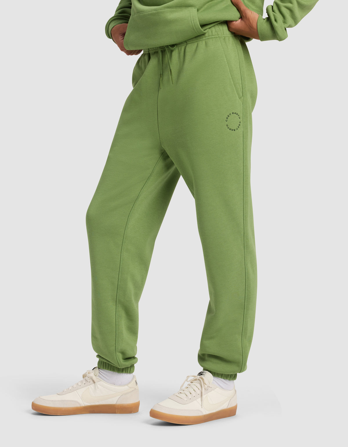 A person wearing Cozy Earth Men's Cityscape Sweatpant in green with a matching sweatshirt, white socks, and beige sneakers stands hands on hips against a plain light background.