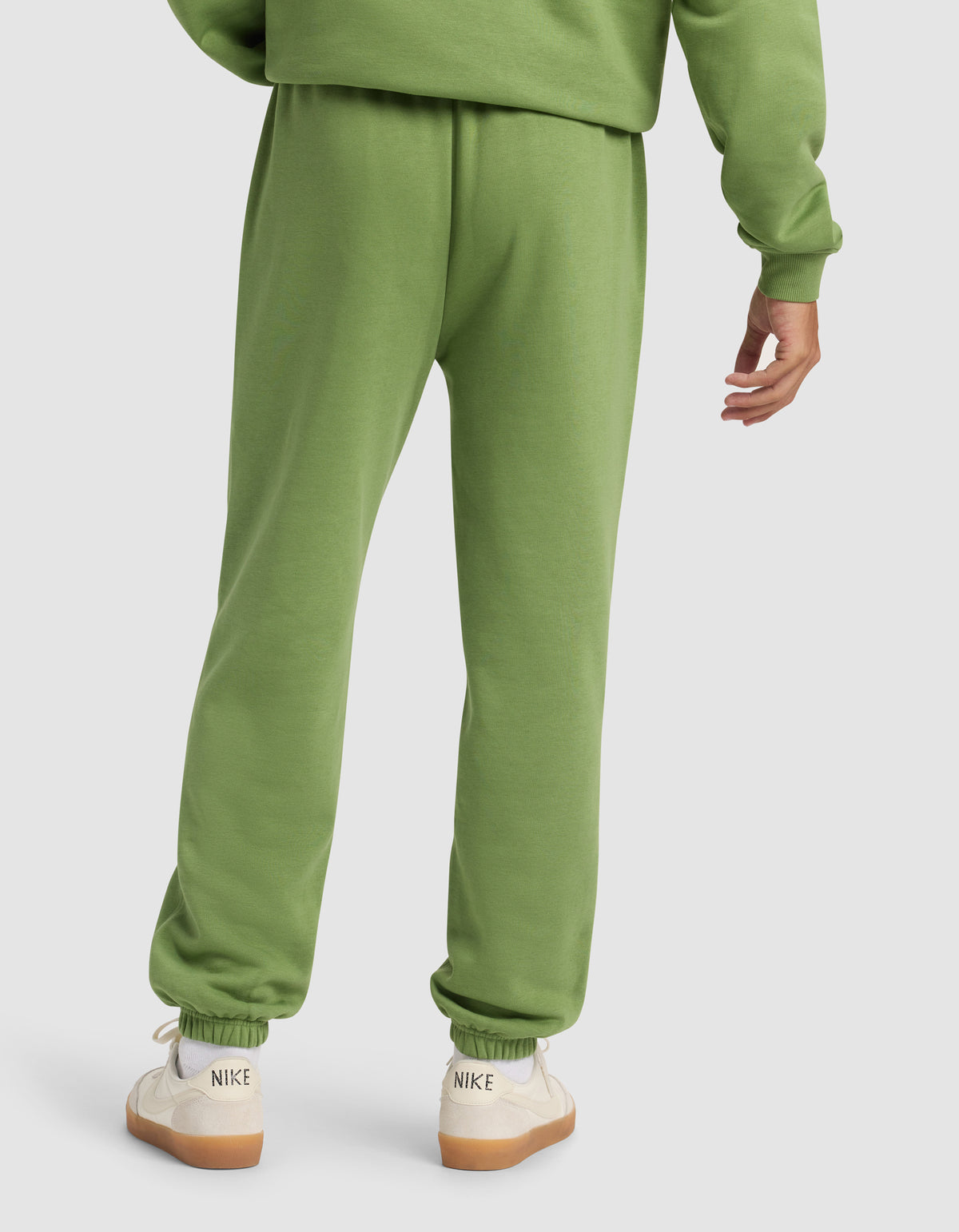 A person is shown from the back wearing Cozy Earth Men's Cityscape Sweatpant and a matching green sweatshirt, paired with white Nike sneakers with beige soles against a plain, light-colored background.