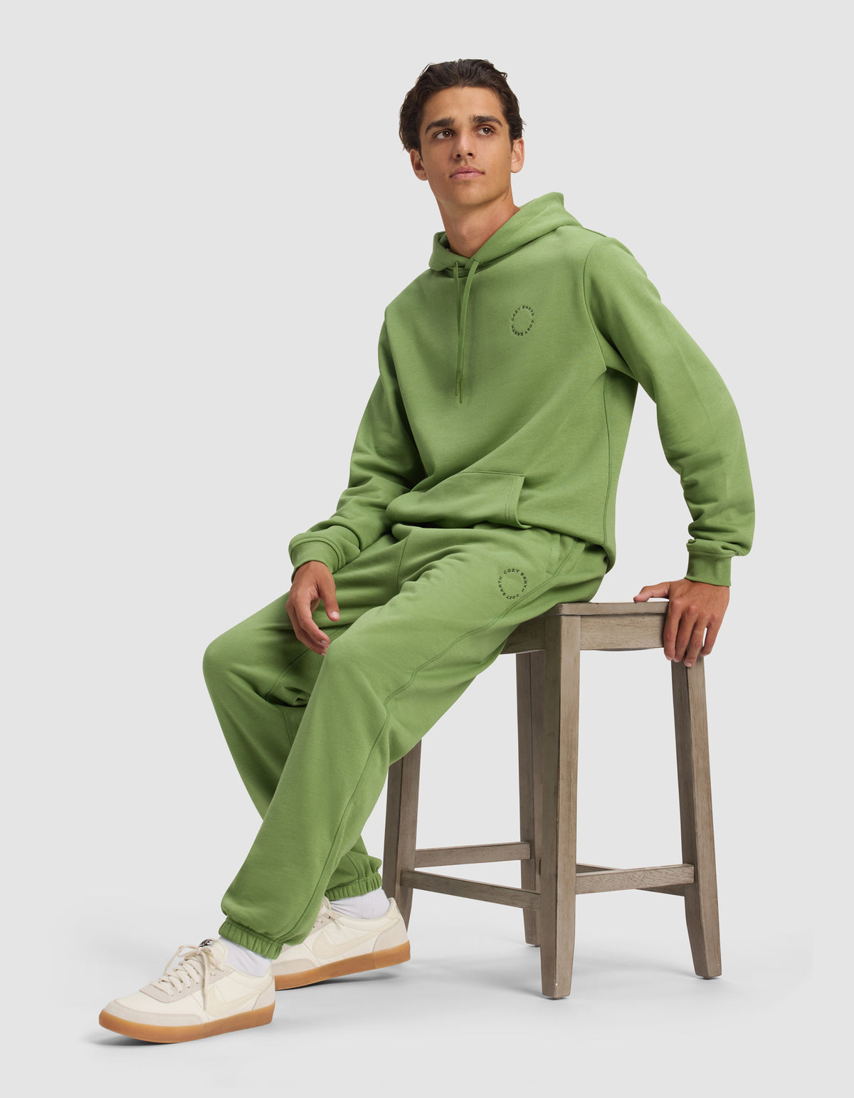 A person in Cozy Earth's Men's Cityscape Sweatpant and a matching light green hoodie sits on a wooden stool, wearing white sneakers with beige soles. They have short brown hair, and the background is plain, light gray.