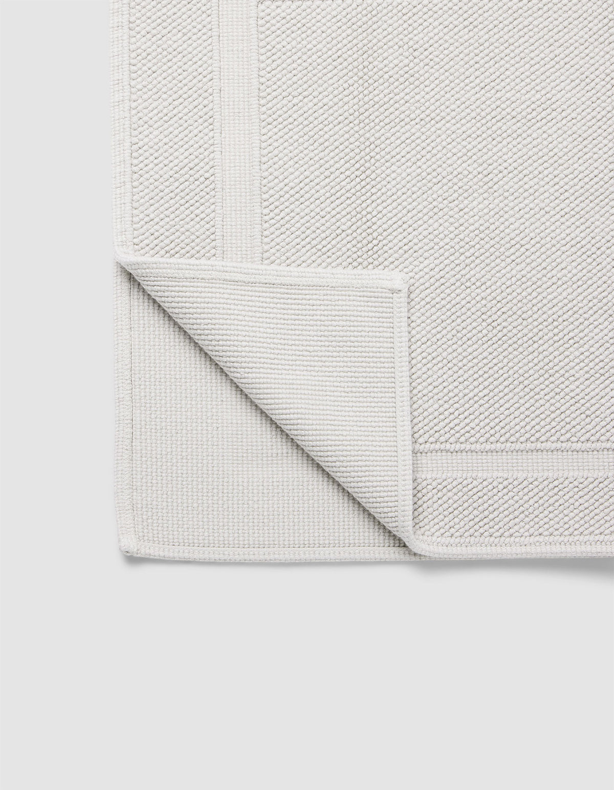 The Cozy Earth Classic Bath Mat in light grey is displayed flat on a plain white background, with one corner folded over to show its woven underside and detailed border.