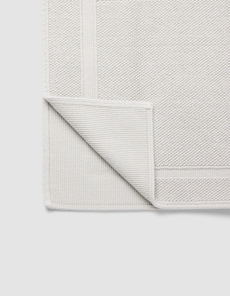 The Cozy Earth Classic Bath Mat in light grey is displayed flat on a plain white background, with one corner folded over to show its woven underside and detailed border.