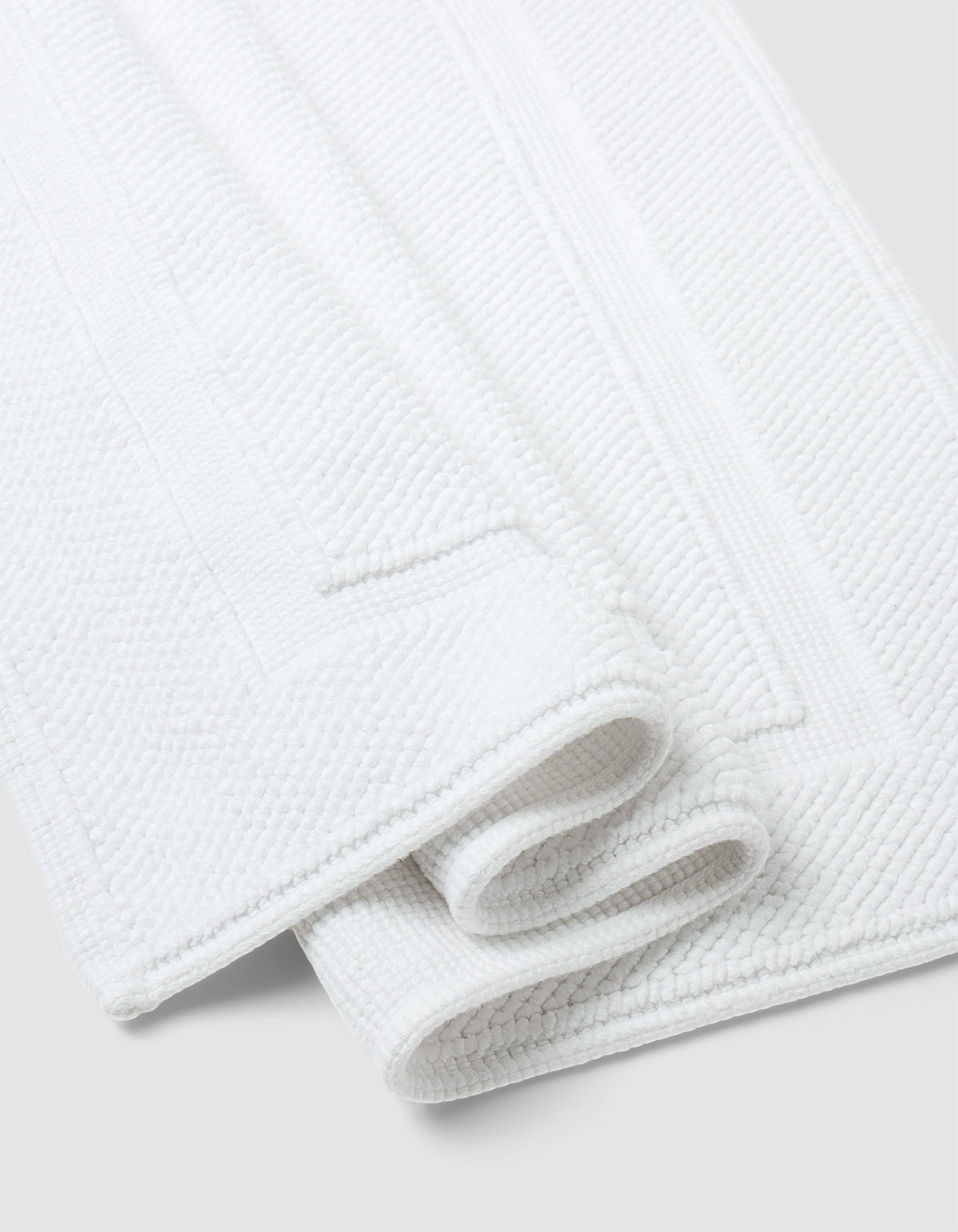 Two neatly folded Cozy Earth Classic Bath Mats, featuring a white textured surface and rectangular border design, are placed on a light gray background.|Color:White