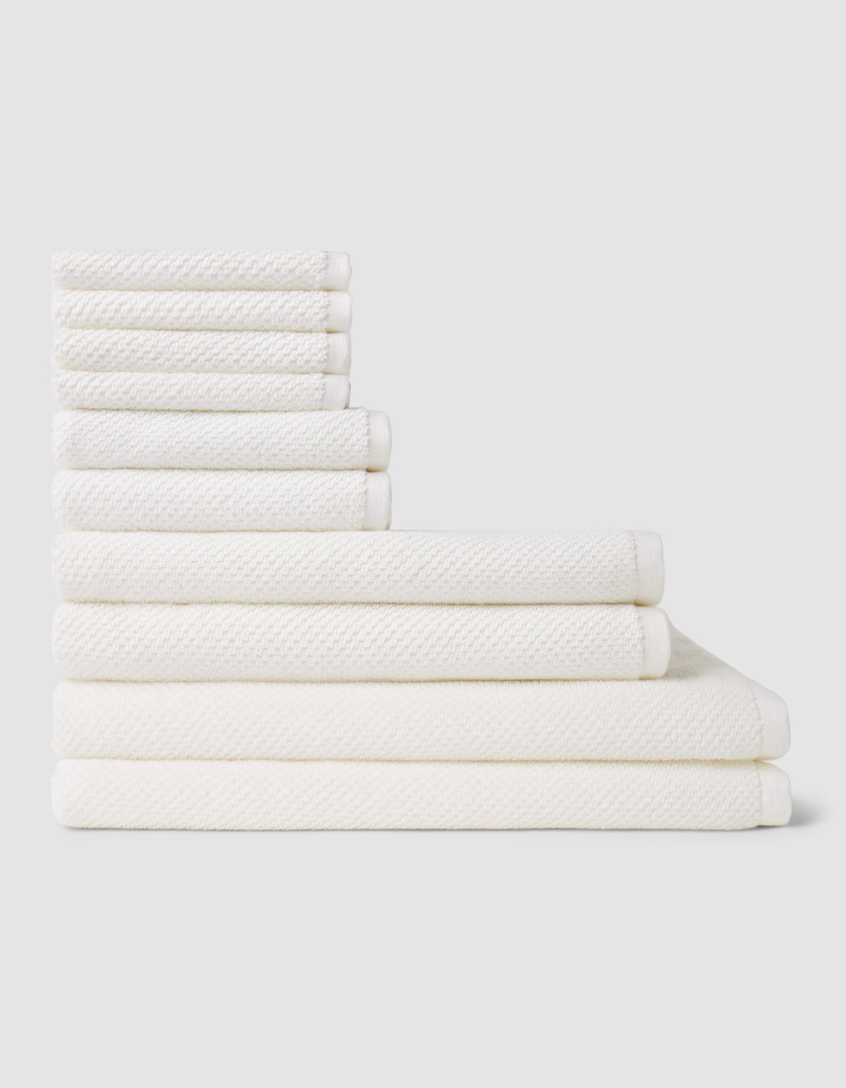 The Nantucket Bath Towel Set by Cozy Earth is shown neatly stacked by size from smallest to largest on a light gray background.|Color:White