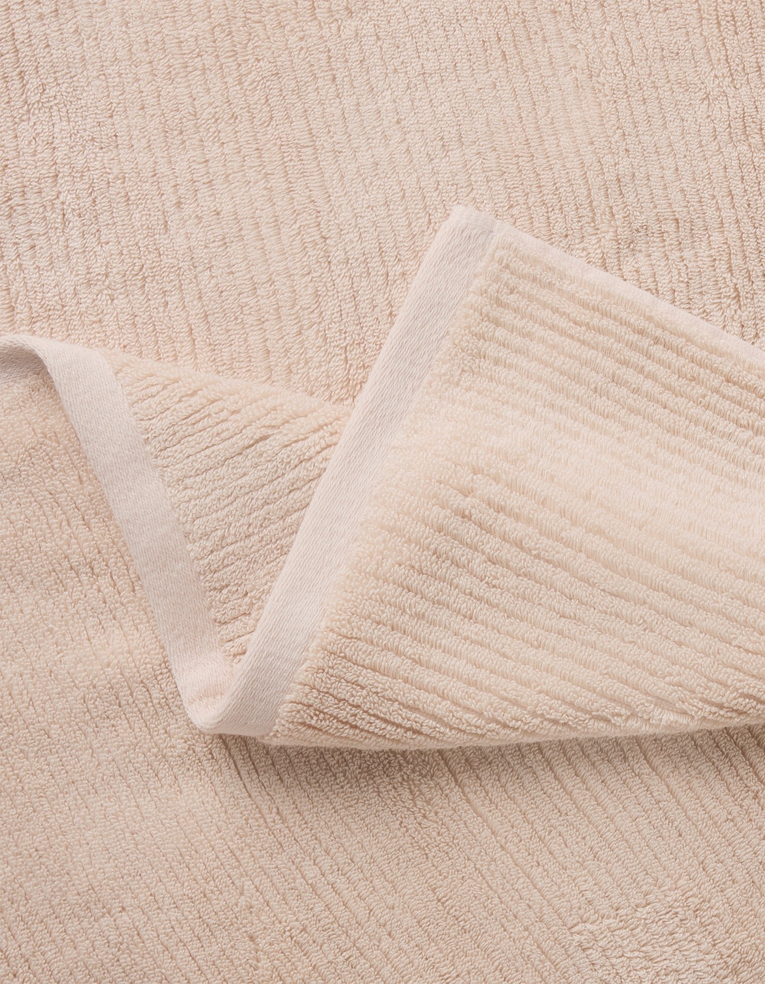A close-up of Cozy Earth’s Complete Ribbed Terry Bath Bundle shows a folded beige towel with a soft, textured surface and subtle horizontal lines resting on a matching background.|Color:Petal