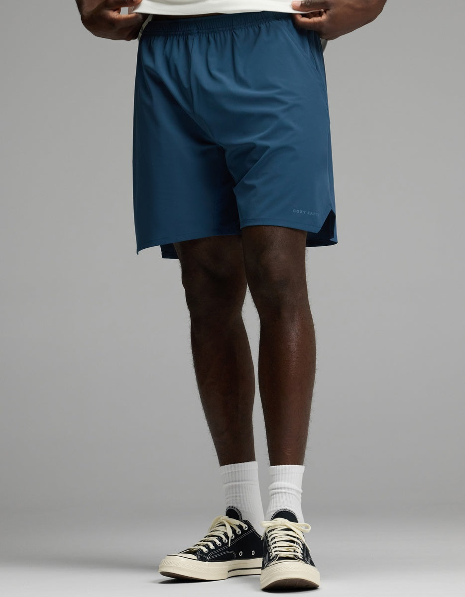 A person is shown from the waist down wearing Cozy Earth’s Men’s Day & Night Short 7″ in Fathom, paired with white socks and black sneakers, standing against a plain gray background.
