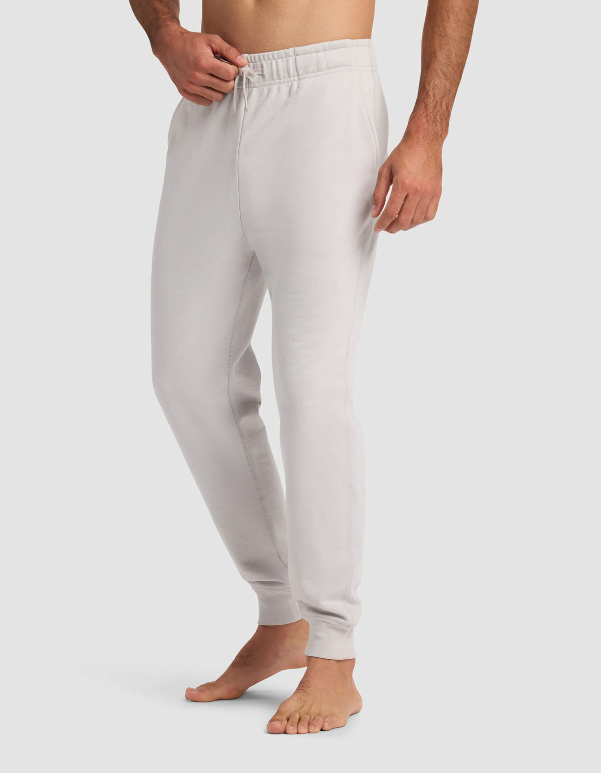 A barefoot man wears Cozy Earth's Men's Dylan Jogger in light gray, featuring an elastic waistband and drawstring, with one hand in his pocket. The background is plain white.