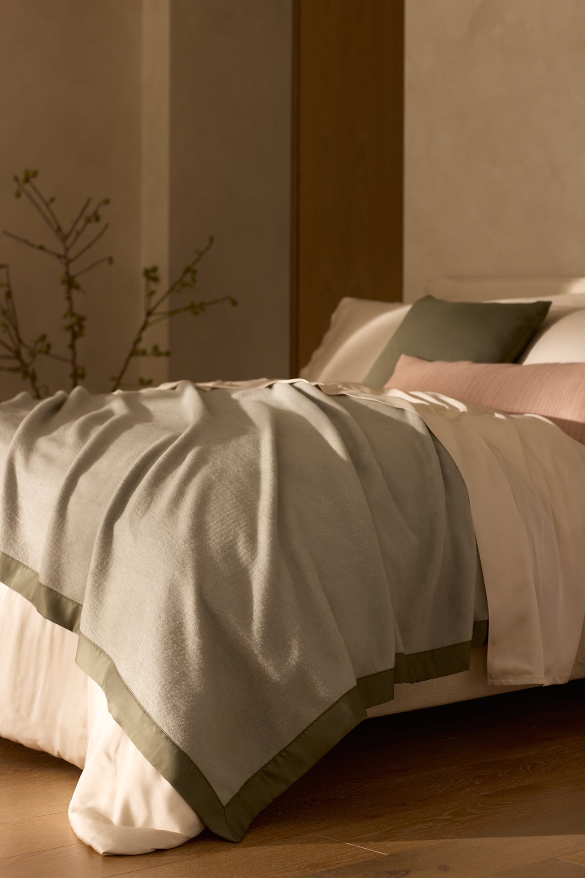 A neatly made bed features the Cozy Earth Bamboo Blanket in light gray with green trim, white sheets, and green and blush accent pillows, set in a softly lit minimalist bedroom with a vase holding a branch nearby. 