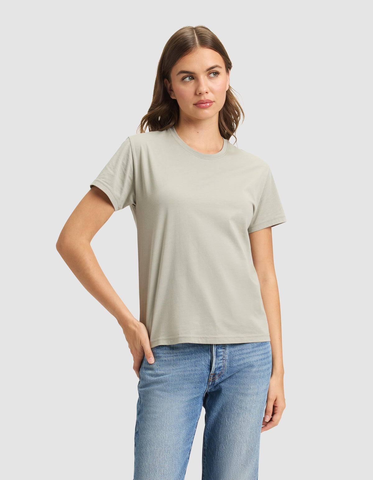 A woman wears the Cozy Earth Women's Easy Tee in light gray with blue jeans, standing against a plain light background, one hand on her hip and a neutral expression. 