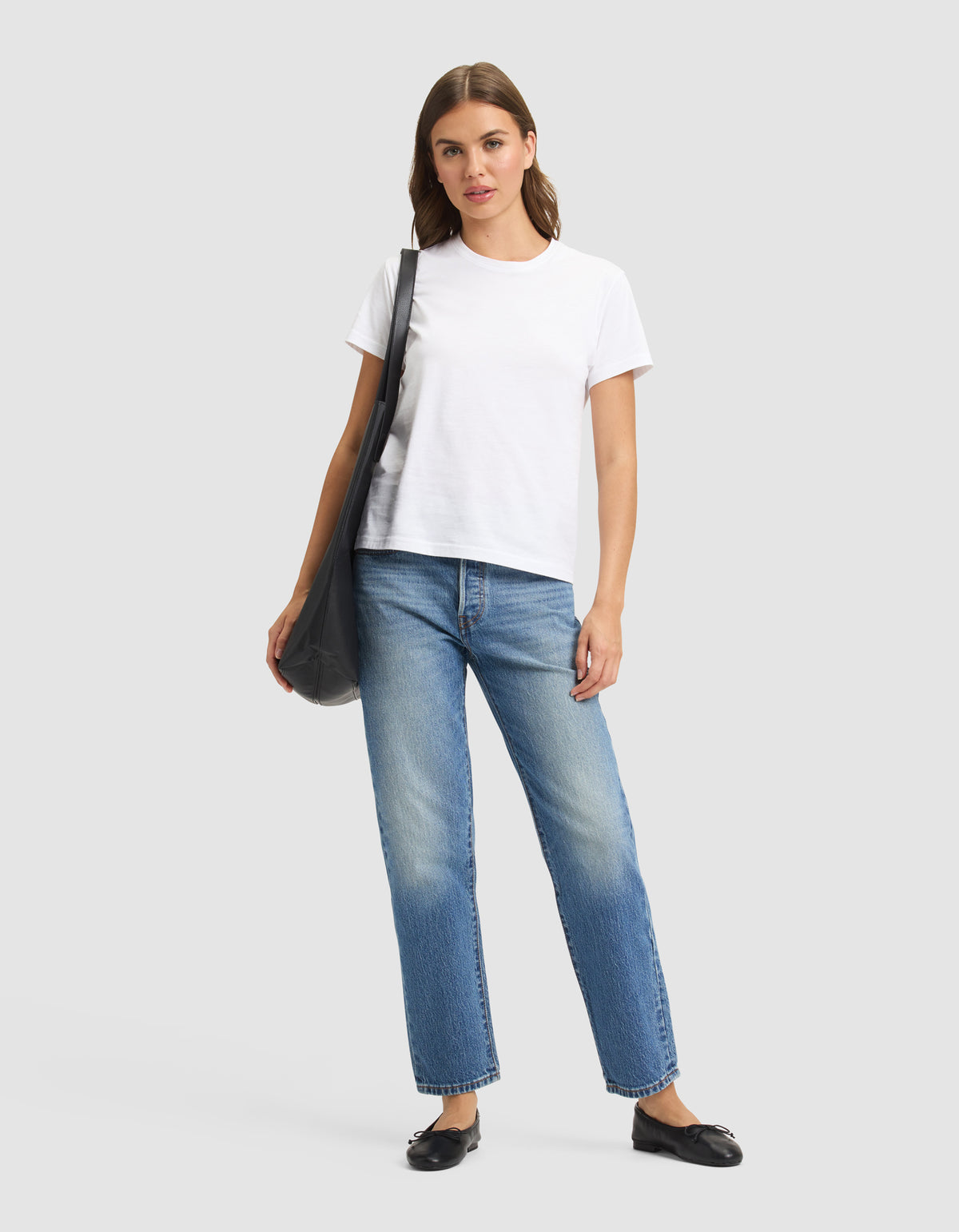 A woman stands against a plain background, wearing the Cozy Earth Women's Easy Tee in white, blue jeans, black flats, and carrying a black shoulder bag. 