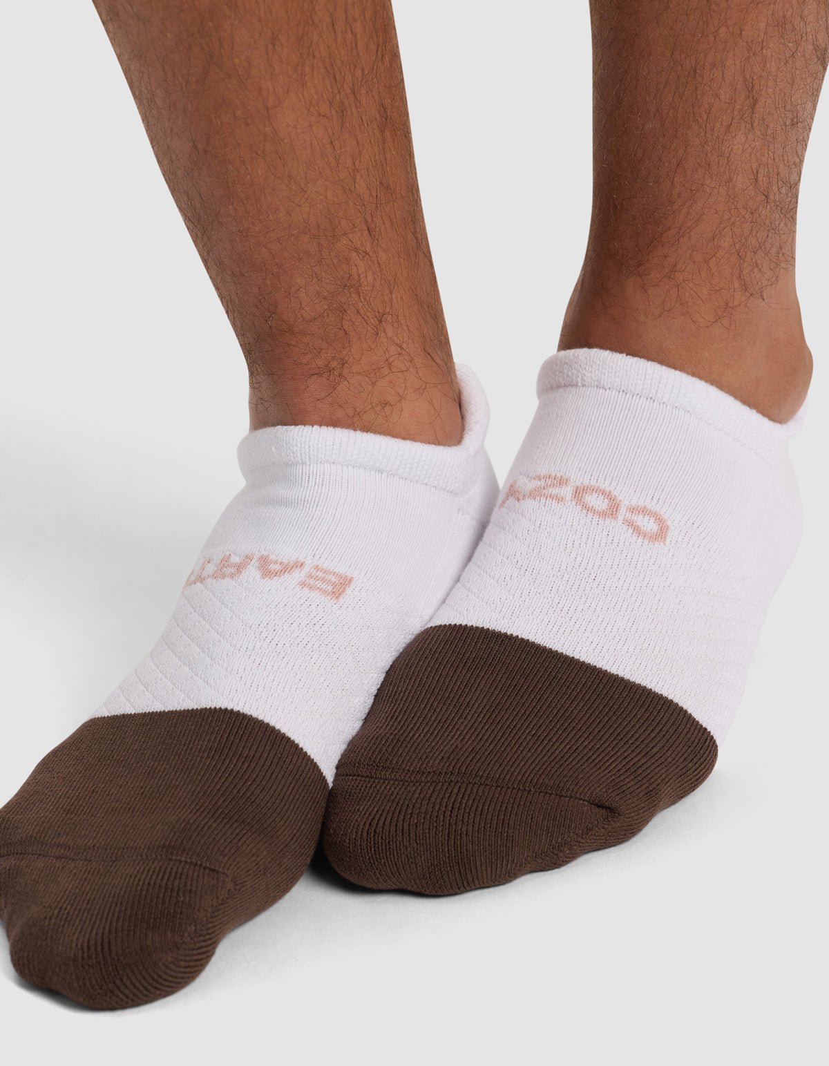 Close-up of feet wearing Cozy Earth Essential Ankle Socks in white with brown toes and faint pinkish text, standing on a light surface.