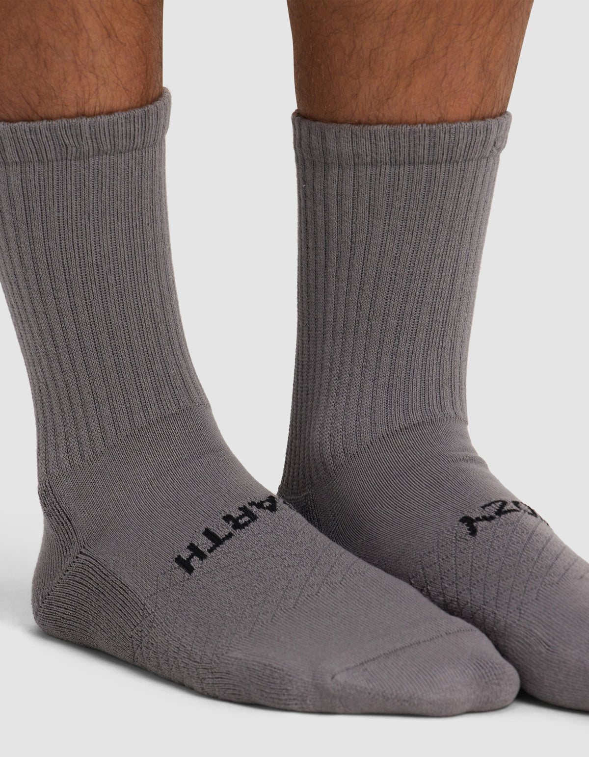 Close-up of a person wearing Cozy Earth Essential Calf Socks in gray with black text near the toes, standing on a white surface. Only the lower legs and feet are visible.