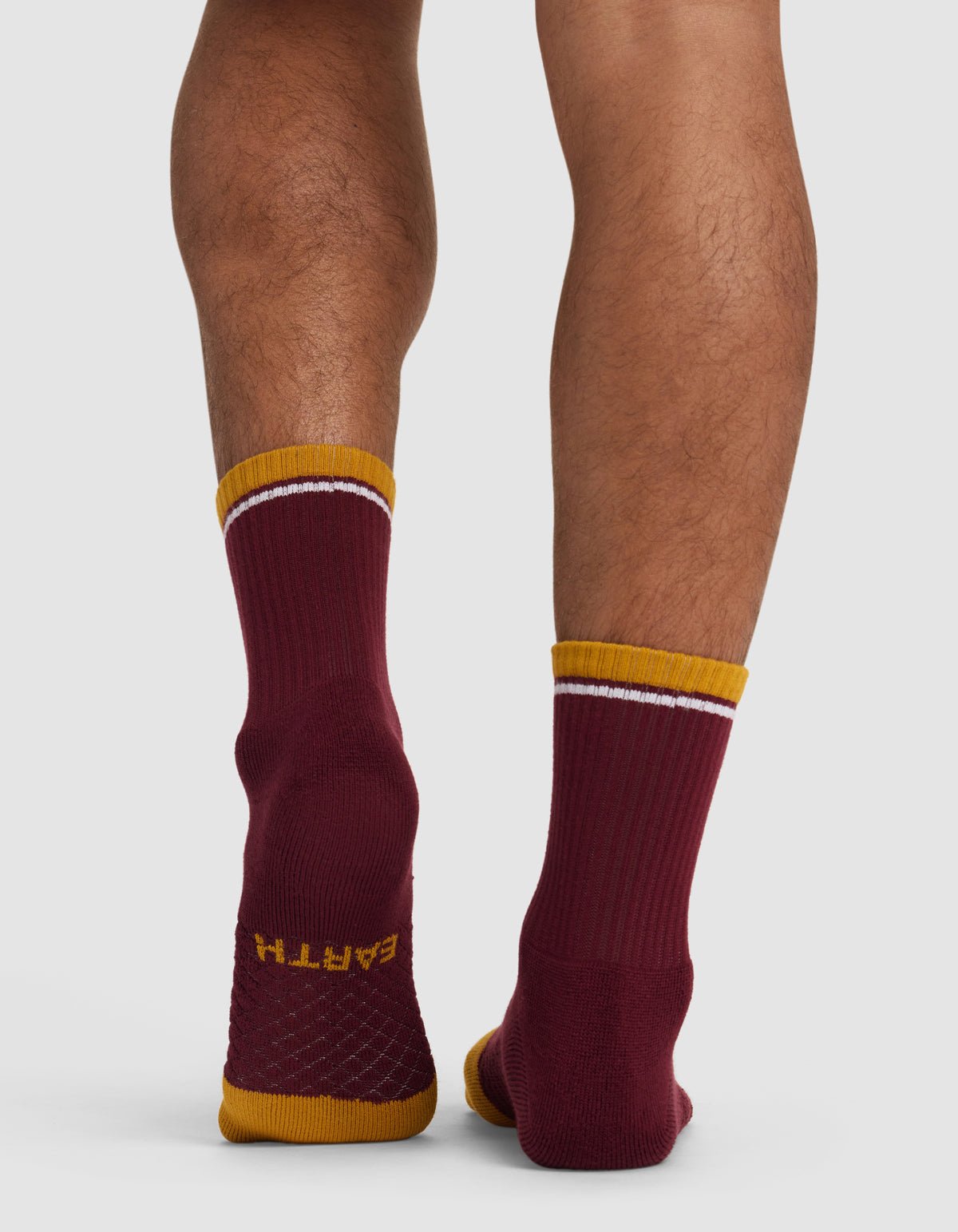 Back view of a person wearing Cozy Earth's Essential Calf Sock—mismatched maroon socks with gold toes and cuffs, white stripes near the top, and "EARTH" on one sole. The person stands on a light background.