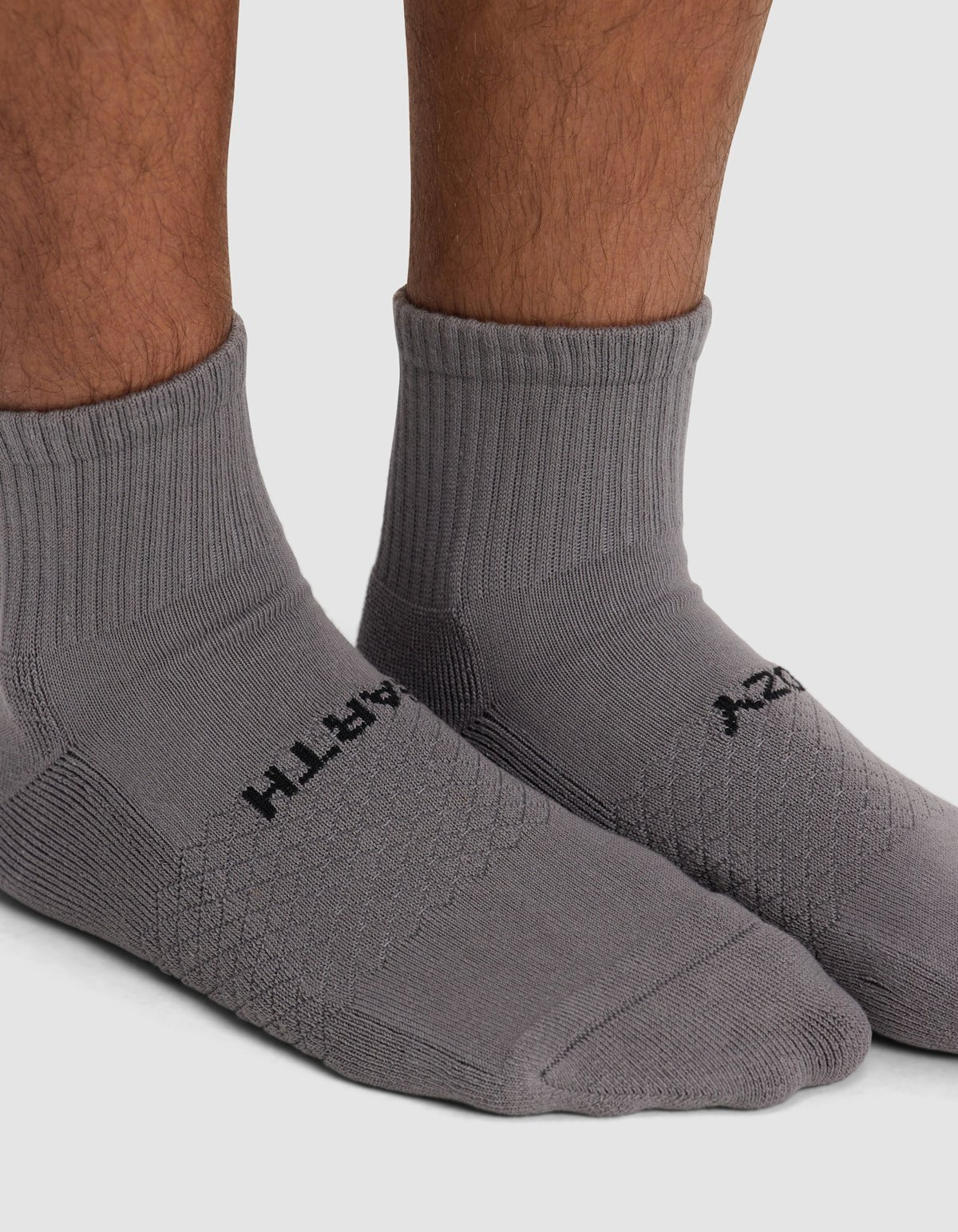 Close-up of a person wearing Cozy Earth's Essential Quarter Sock in gray with black text, standing on a white background.
