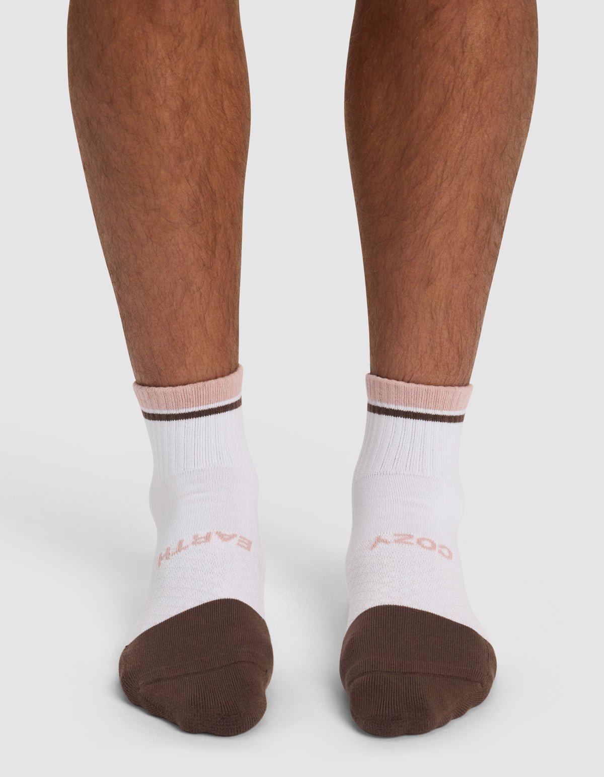 A person wears Cozy Earth's Essential Quarter Sock (white with brown toes and stripe, "EARTH" in light pink on left sock) while standing on a white background.