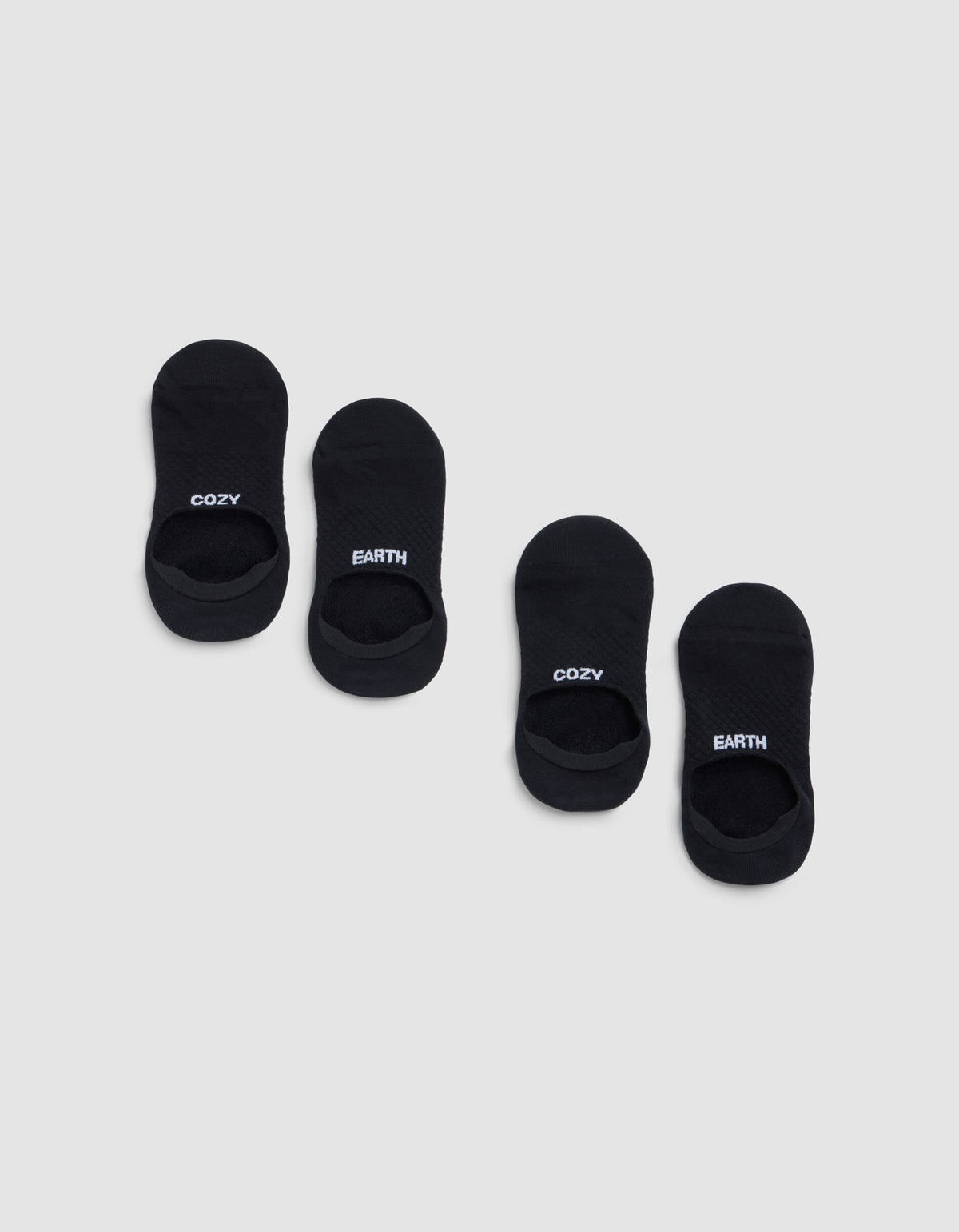 Two pairs of black Essential No-Show Socks by Cozy Earth are displayed on a light gray background. One pair features "COZY" in white lettering, and the other pair displays "EARTH" in white. 
