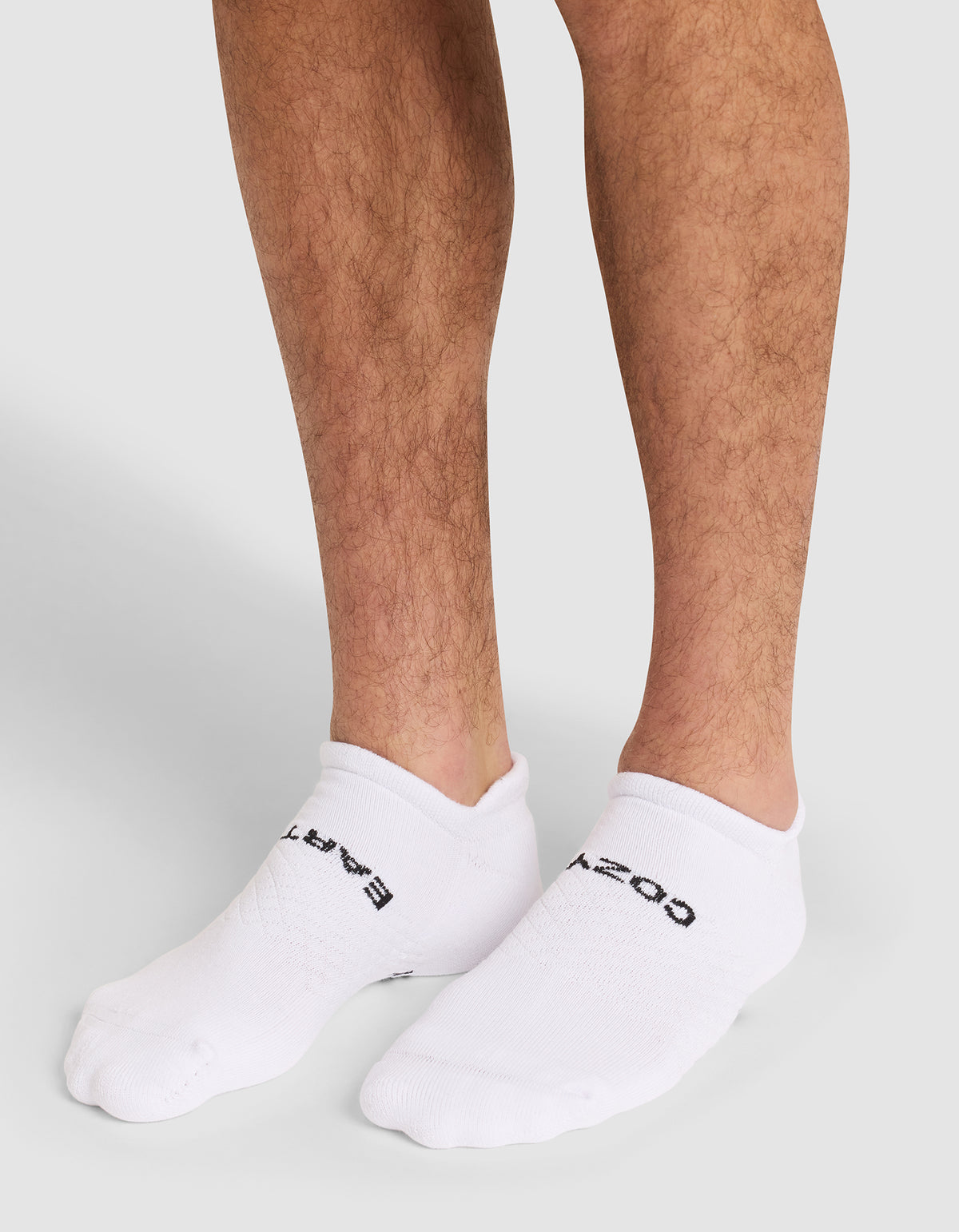 A close-up of a person's lower legs wearing Cozy Earth Essential Ankle Socks in white with black text, standing on a neutral gray background. 