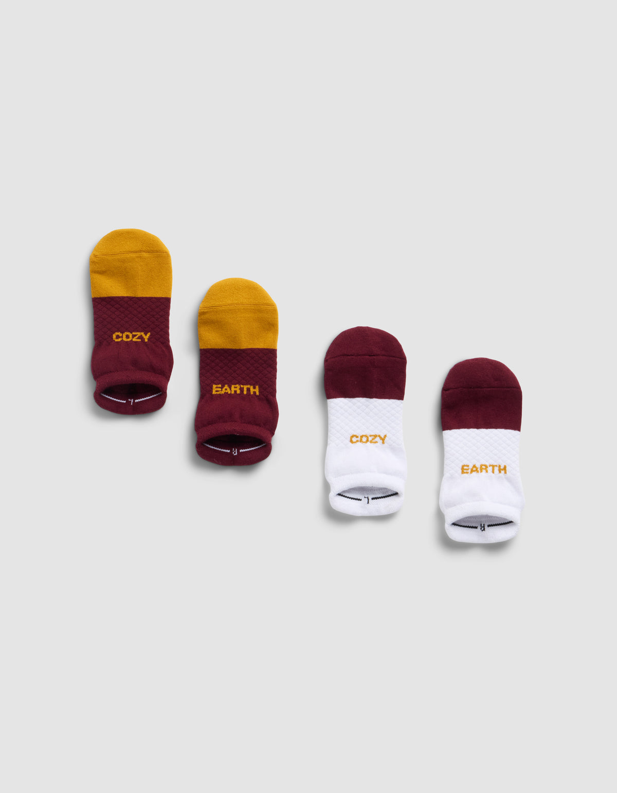 Four pairs of Cozy Earth Essential Ankle Socks on a gray background: the left pair is yellow and burgundy with "COZY" and "EARTH" text, the right pair is burgundy and white with the same words on each sock.