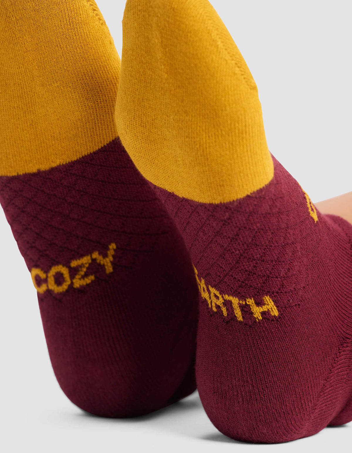 Close-up of two feet wearing Cozy Earth Women's Essential Ankle Socks featuring maroon heels, yellow tops, and "COZY" and "EARTH" in yellow text on each heel. The background is plain light gray.