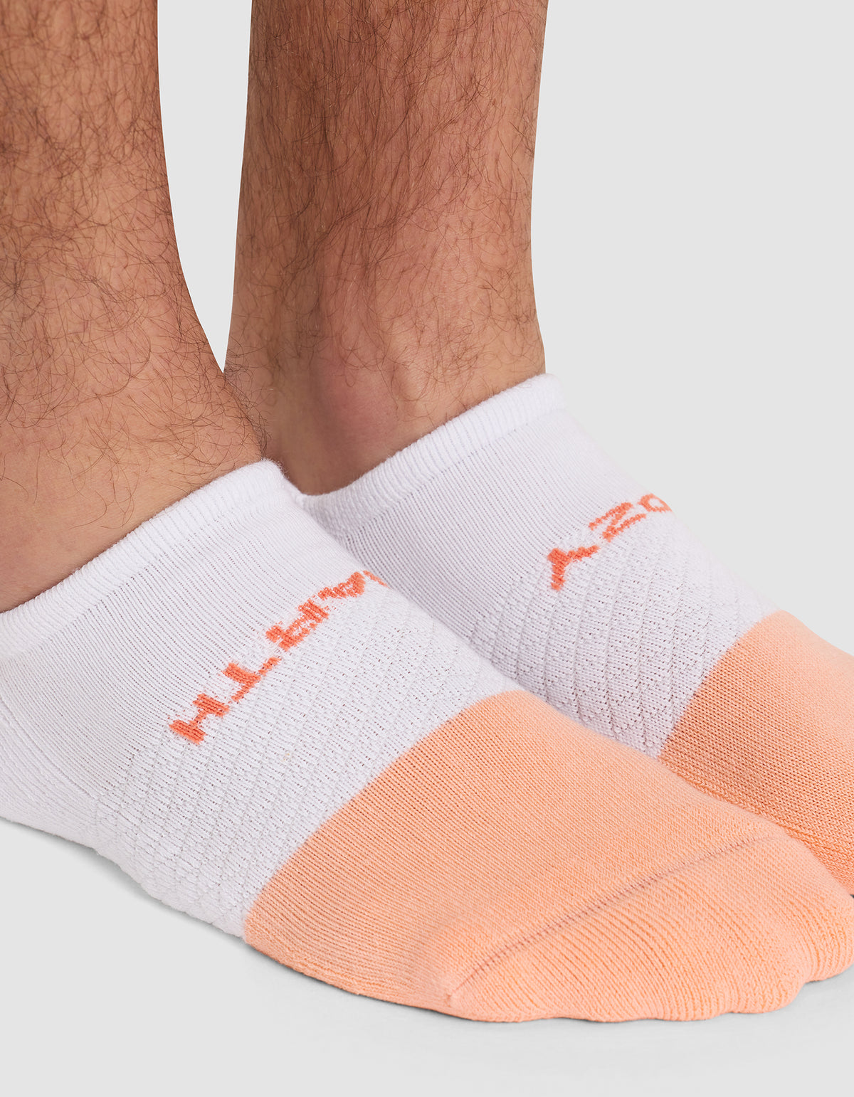 Close-up of a person's lower legs and feet wearing Cozy Earth Essential Ankle Socks in white and light orange, featuring orange text on top. Background is plain light gray. 