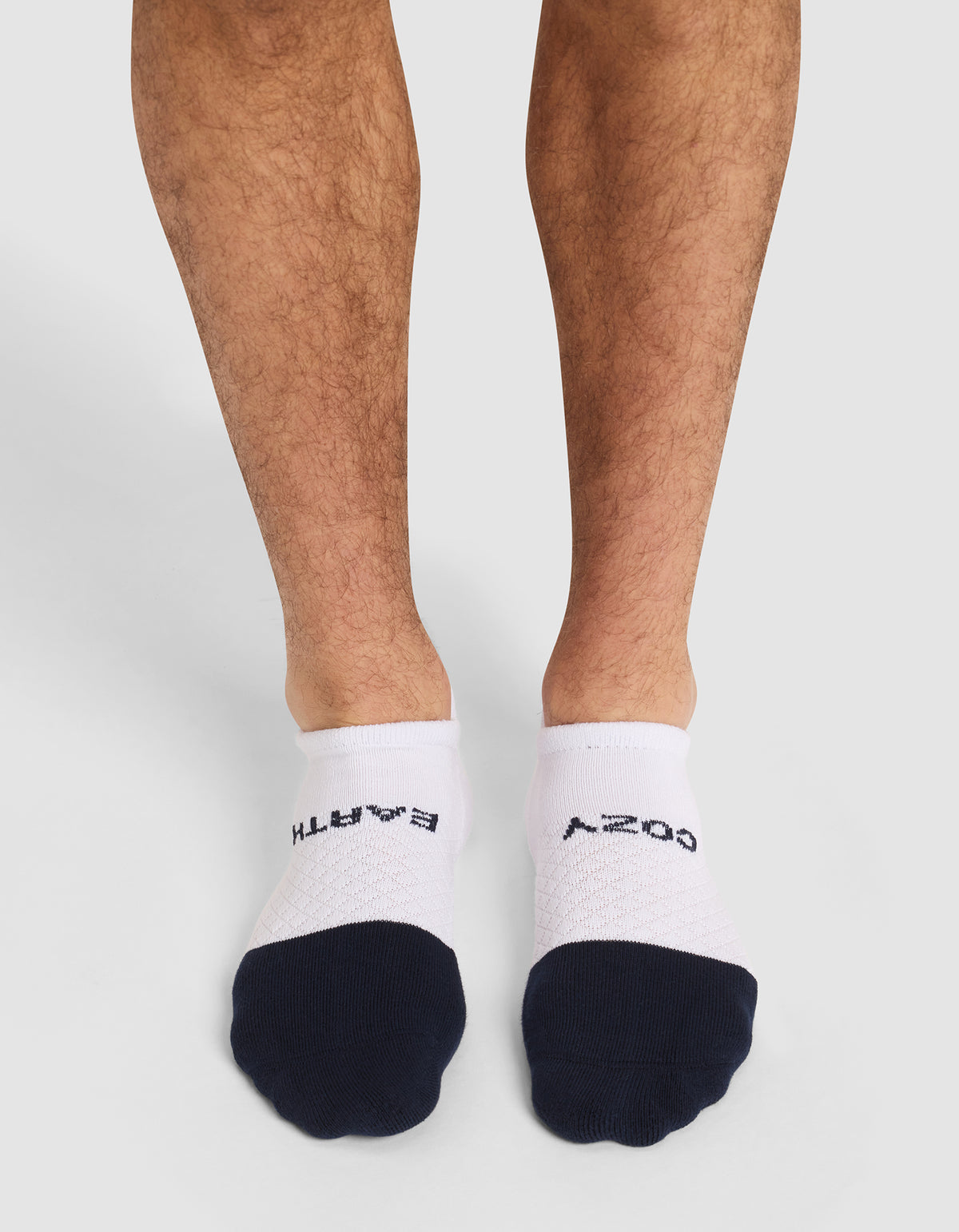 A person stands on a white background wearing Cozy Earth Essential Ankle Socks—white no-show socks with black toes; "BATH" is written on the left sock and "COZY" on the right in black letters. 