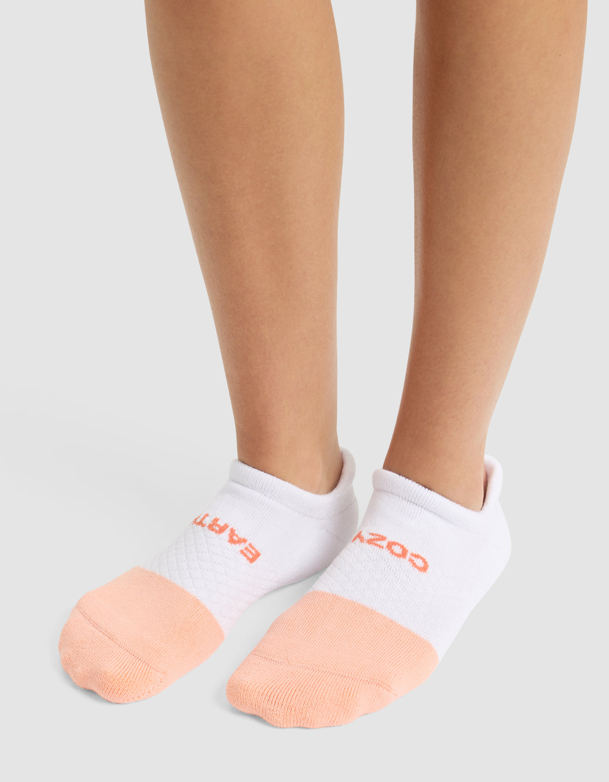 A person wearing Cozy Earth's Essential Ankle Sock—featuring white tops, peach toes, and red text near the cuff—stands on a light gray surface. 