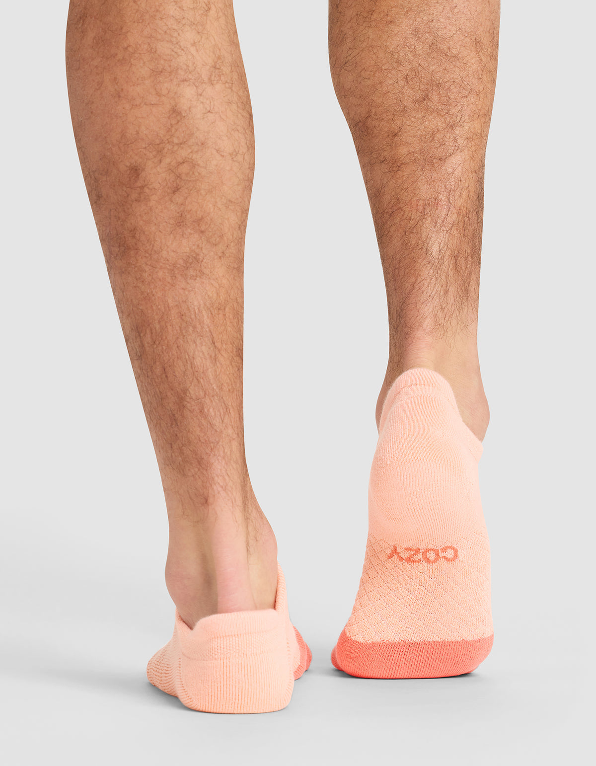 A person with hairy legs is standing on a light surface, wearing peach-colored Cozy Earth Essential Ankle Socks that cover the feet up to the ankles, with "COZY" visible on one heel. 