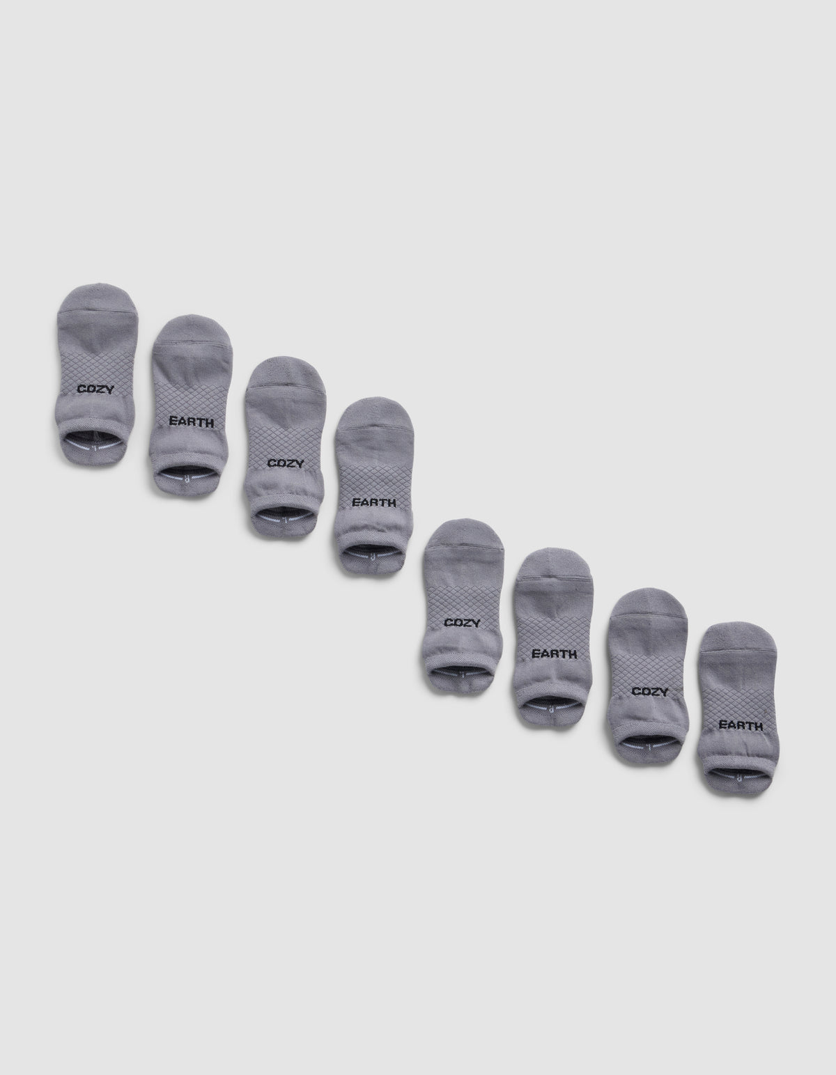 Four pairs of gray Essential Ankle Socks from Cozy Earth are arranged diagonally on a light gray background, each featuring "COZY EARTH" printed in black near the toes.|Color:Stone