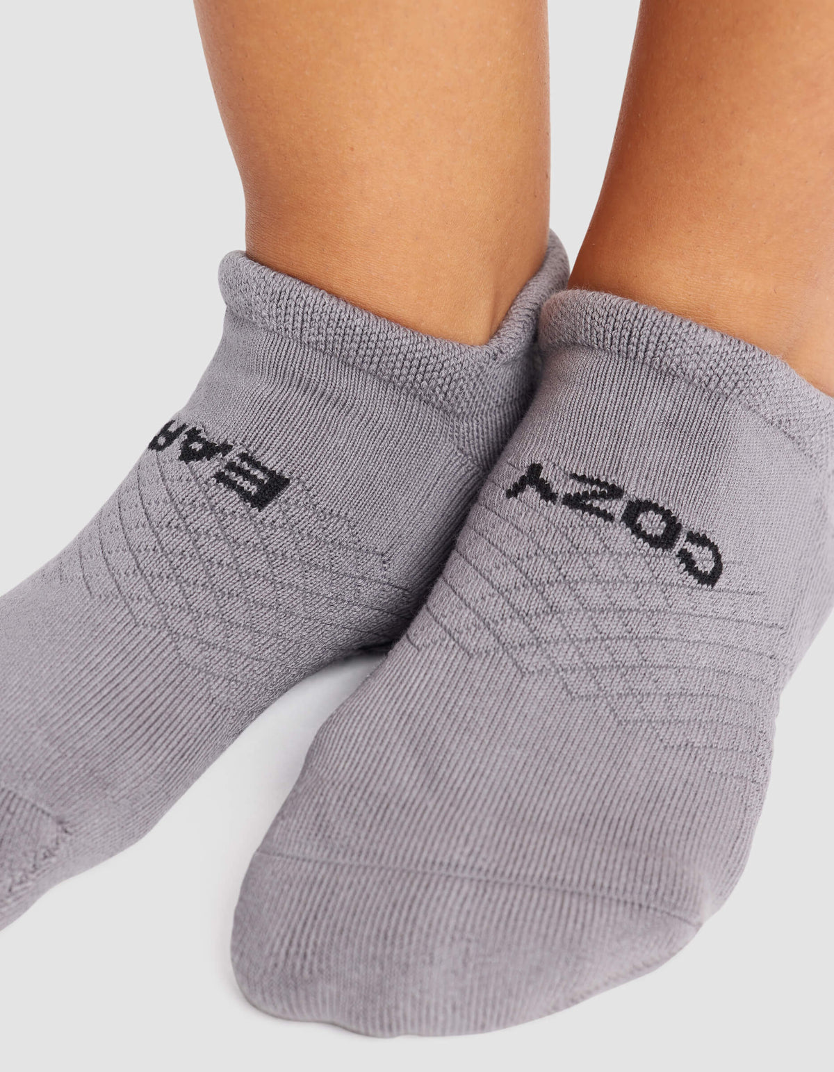 Close-up of feet wearing Cozy Earth Women's Essential Ankle Socks in light gray, featuring black "COZY" on one sock and "BARE" on the other, both clearly visible against a neutral background.