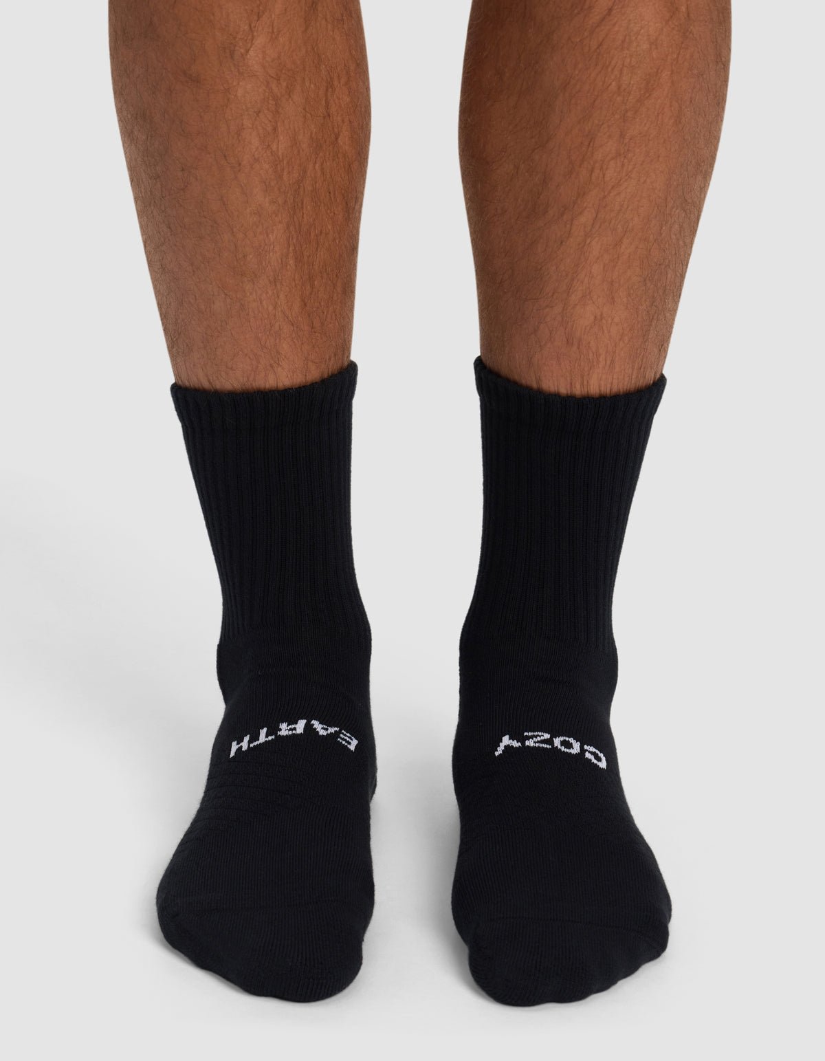 A person wears Cozy Earth Essential Calf Socks in black, featuring "EARTH" on the left and "COZY" on the right sock. Only their lower legs and feet appear against a plain background.