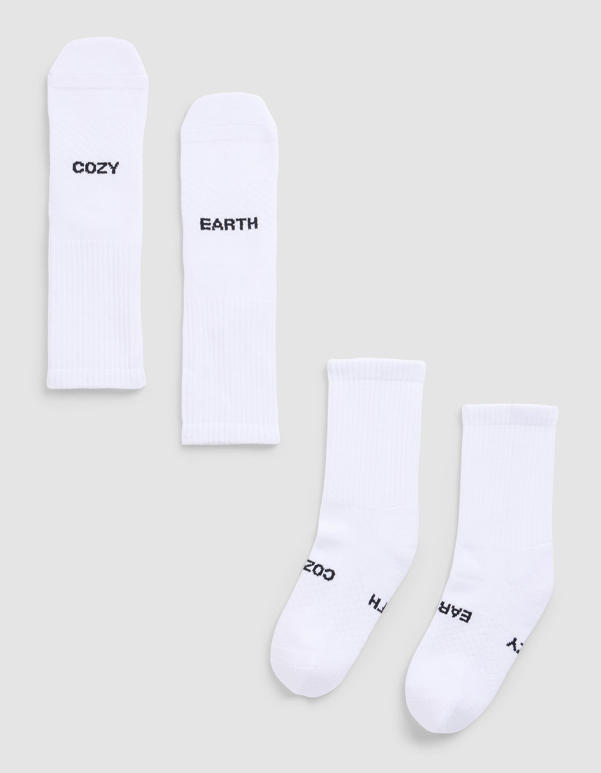 Four Essential Calf Socks by Cozy Earth are displayed on a light gray surface. Each white ribbed sock features black text: two say "COZY," and two say "EARTH." The socks are arranged flat and side by side. 