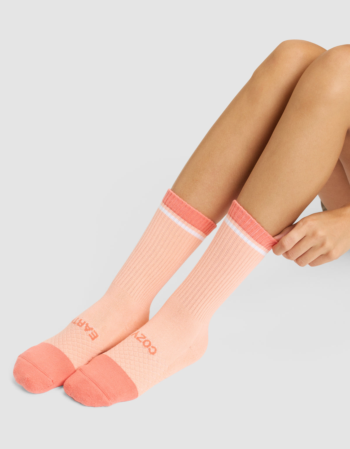 A person sits on the floor wearing Essential Calf Socks by Cozy Earth—peach crew socks with coral toes, heels, and cuffs, a white stripe near the top, and text on the toes. Only their legs and hands are visible. 