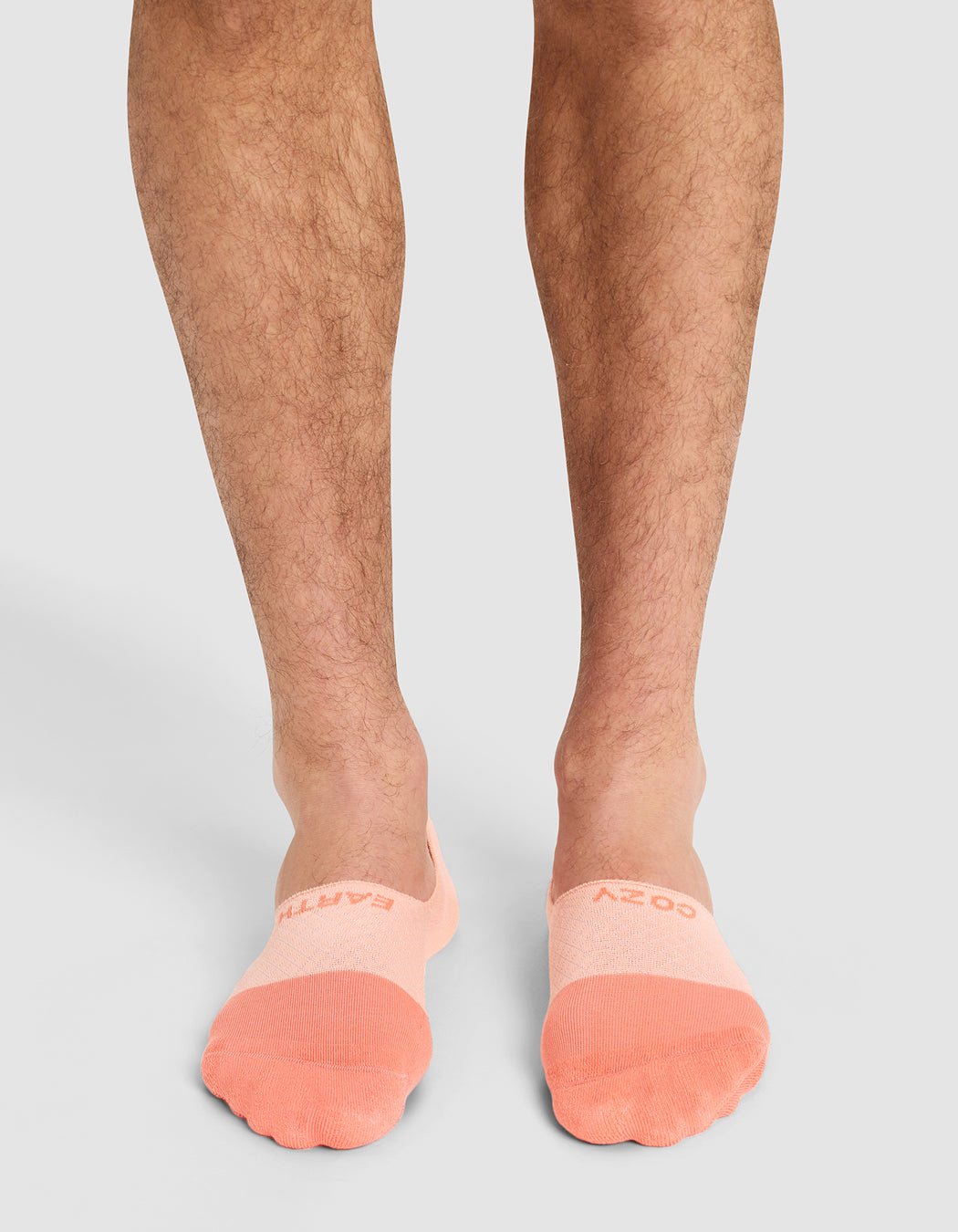 A person wearing Cozy Earth Essential No-Show Socks in light pink with peach toes, featuring text near the toes, stands on a light gray surface. Only their lower legs and feet are visible.