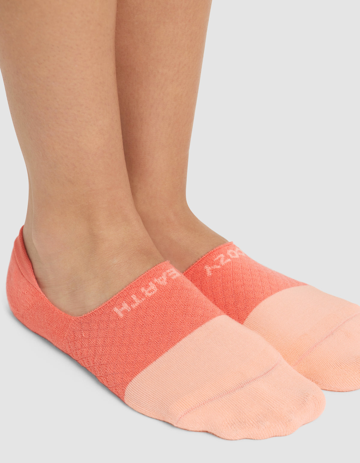 A close-up of two feet wearing Cozy Earth’s Essential No-Show Socks in peach and coral, featuring partially visible white "CRAZY EARTH" text near the ankle, standing on a light gray surface. 