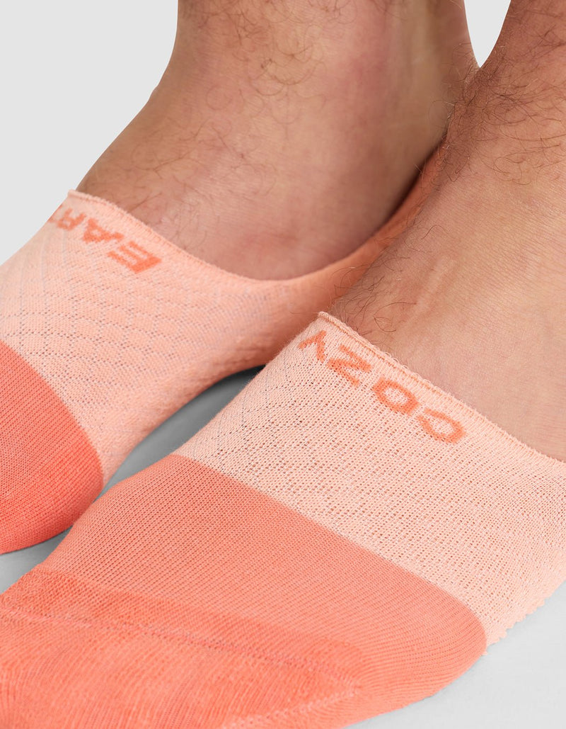 Close-up of two feet wearing Cozy Earth Essential No-Show Socks in light peach and coral with subtle texture. "COZY" is visible on one sock. Background is plain, light gray.